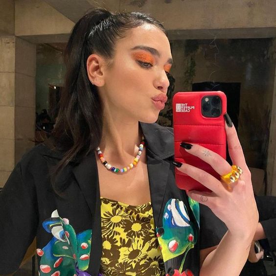 A woman with long dark hair in a high ponytail takes a mirror selfie. She wears bright orange eyeshadow, a colorful beaded necklace, a black shirt with floral prints, and holds a red phone case.