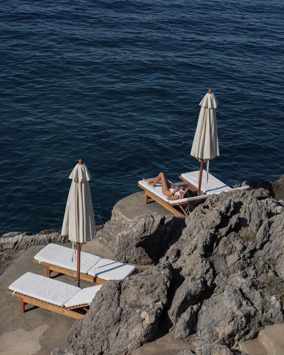 A person relaxes on a sun lounger under a closed umbrella on rocky seaside terrain, with another empty lounger nearby. The calm, deep blue ocean stretches out in the background.