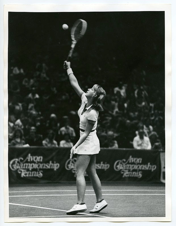 A female tennis player in mid-serve prepares to hit the ball on an indoor court, with a crowd watching in the background and Avon Championship Tennis signs visible. The image is in black and white.