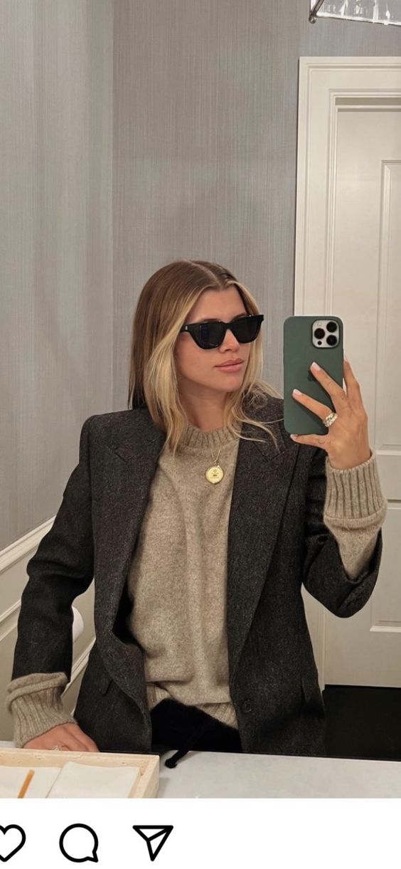 A woman wearing sunglasses, a gray blazer, and a beige sweater takes a mirror selfie with her smartphone in a modern bathroom. She has blonde hair and is posing with a neutral expression.
