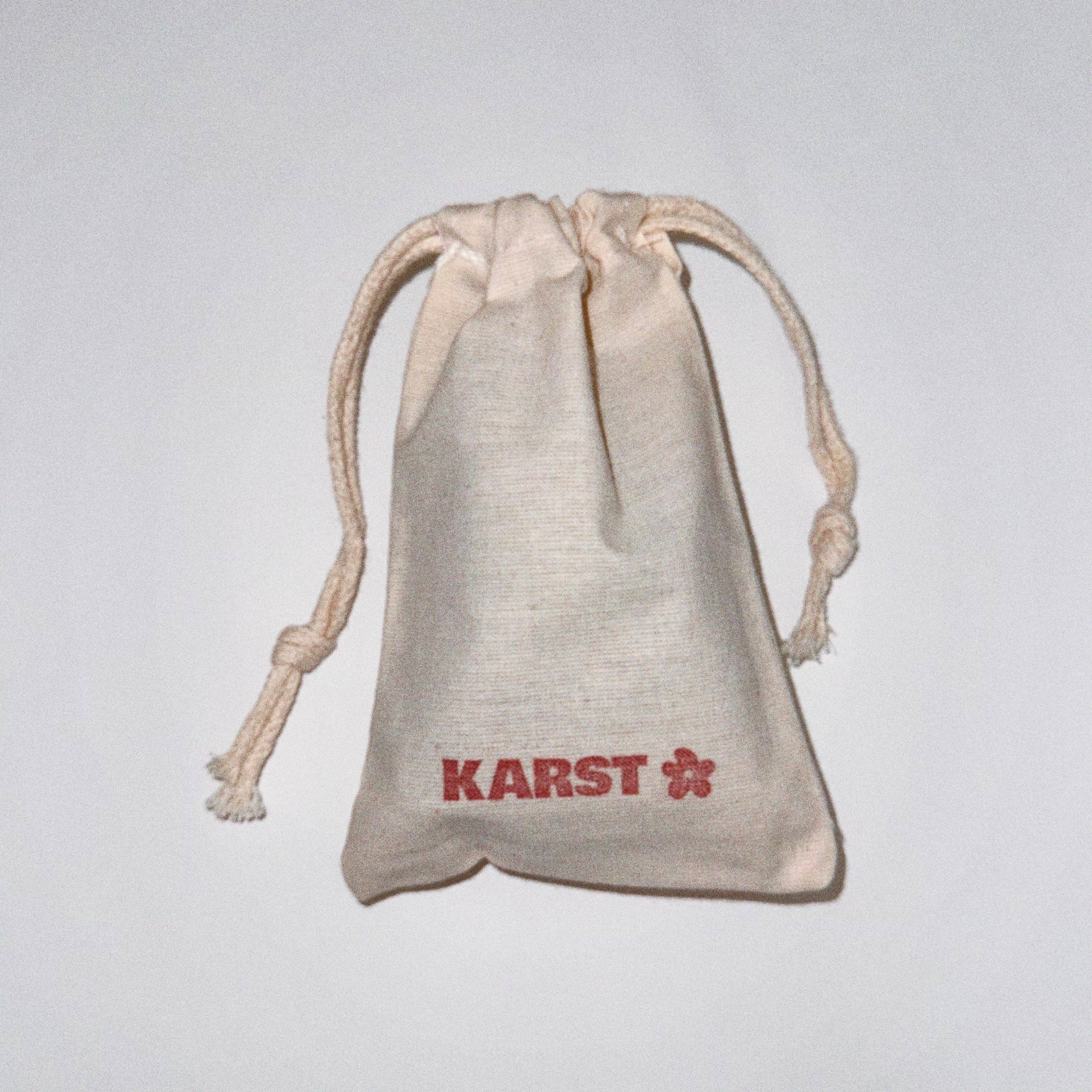 A small beige drawstring pouch with the Karst Essentials logo and flower printed in red, ideal for storing your SuperClip Hair Claw — Maraschino: No Charm Eyelet (Standard). Displayed on a plain white background.