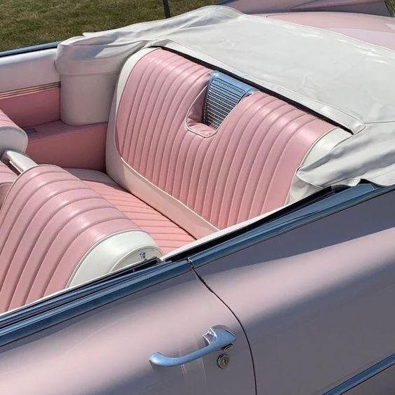 A close-up of a vintage convertible car with pastel pink and white leather seats, a partially folded white convertible top, and chrome detailing on the back seat. The car is parked on grass.
