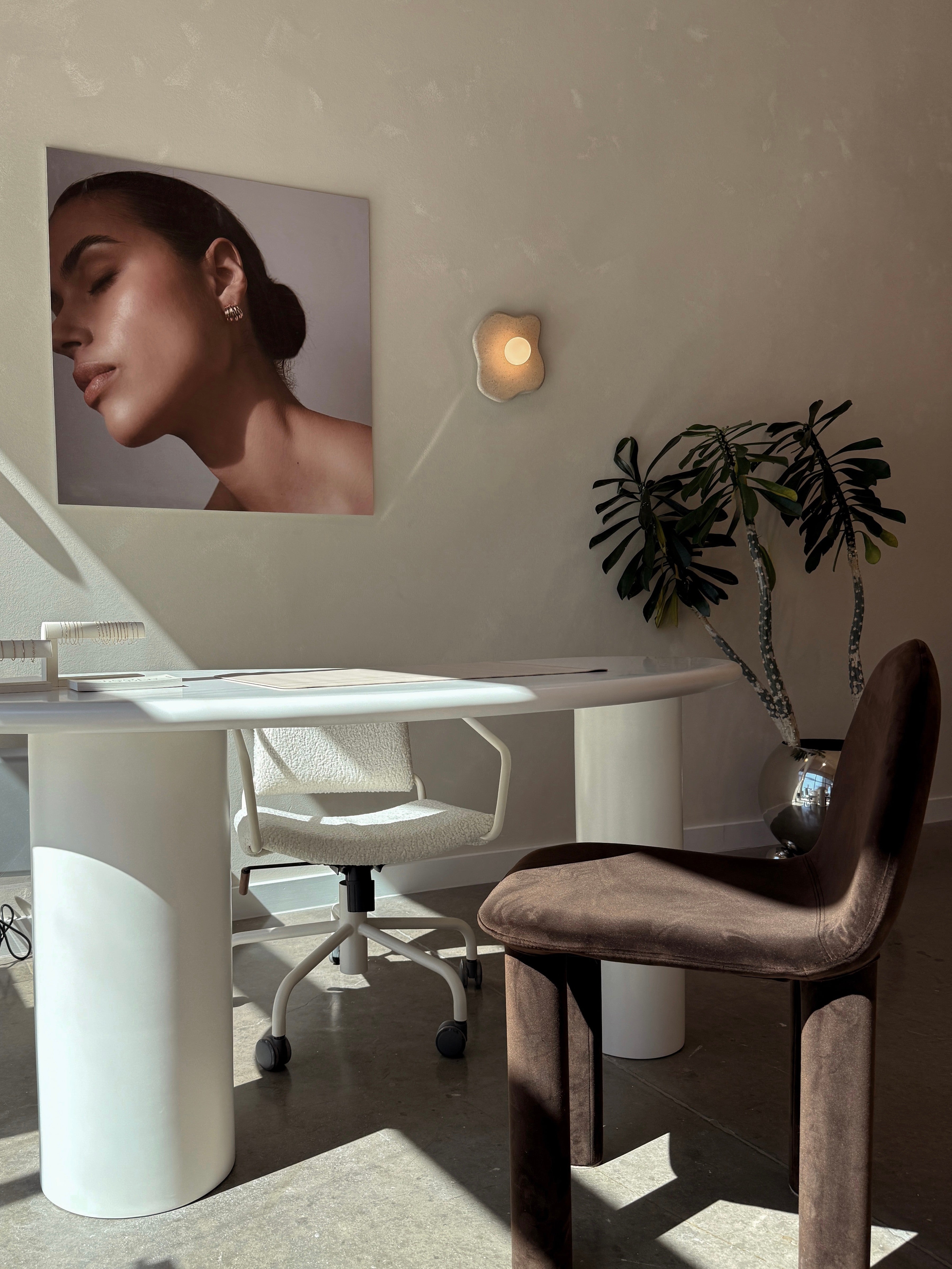 A modern office with a white desk, white office chair, and brown velvet chair. A large portrait of a woman’s face and a potted plant decorate the minimalist, sunlit space.