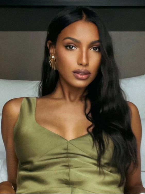 A woman with long, wavy dark hair wearing a gold earring and a sleeveless olive green top sits against a white pillow, looking directly at the camera with a calm expression.