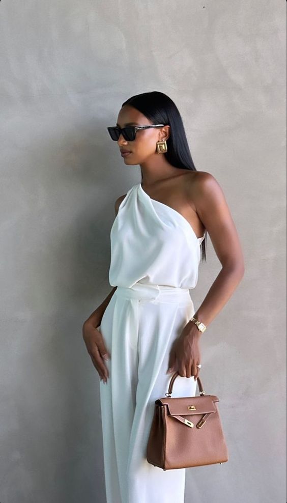 A woman wearing a stylish white one-shoulder top and matching pants poses against a neutral wall. She accessorizes with black sunglasses, gold earrings, a watch, and holds a small brown handbag.