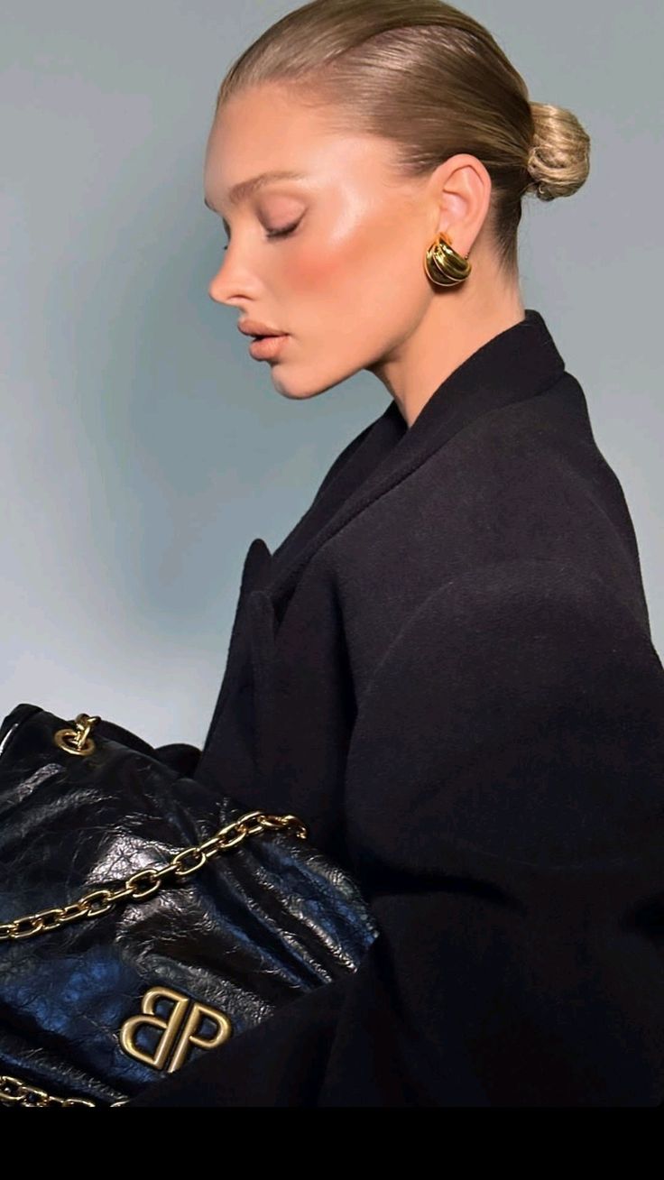 A woman with slicked-back hair in a bun wears a black coat and gold hoop earrings, holding a black leather bag with a gold chain and BP logo, posing in profile against a neutral background.