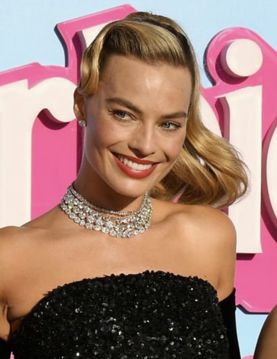 A woman with wavy blonde hair and red lipstick smiles while wearing a strapless black sequin dress, diamond choker necklaces, and stud earrings, standing in front of a colorful backdrop.