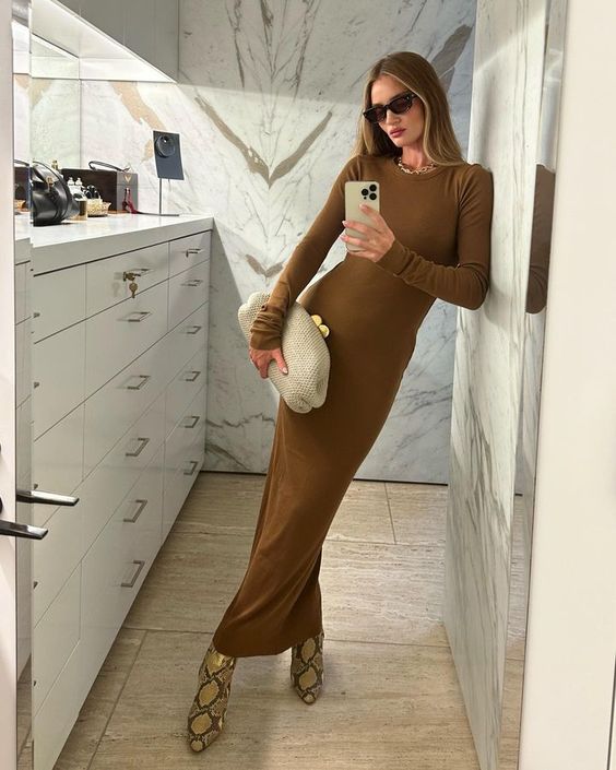 A woman wearing a long brown dress, snakeskin boots, and sunglasses takes a mirror selfie in a modern bathroom with marble walls and light wood floors, holding a beige clutch.