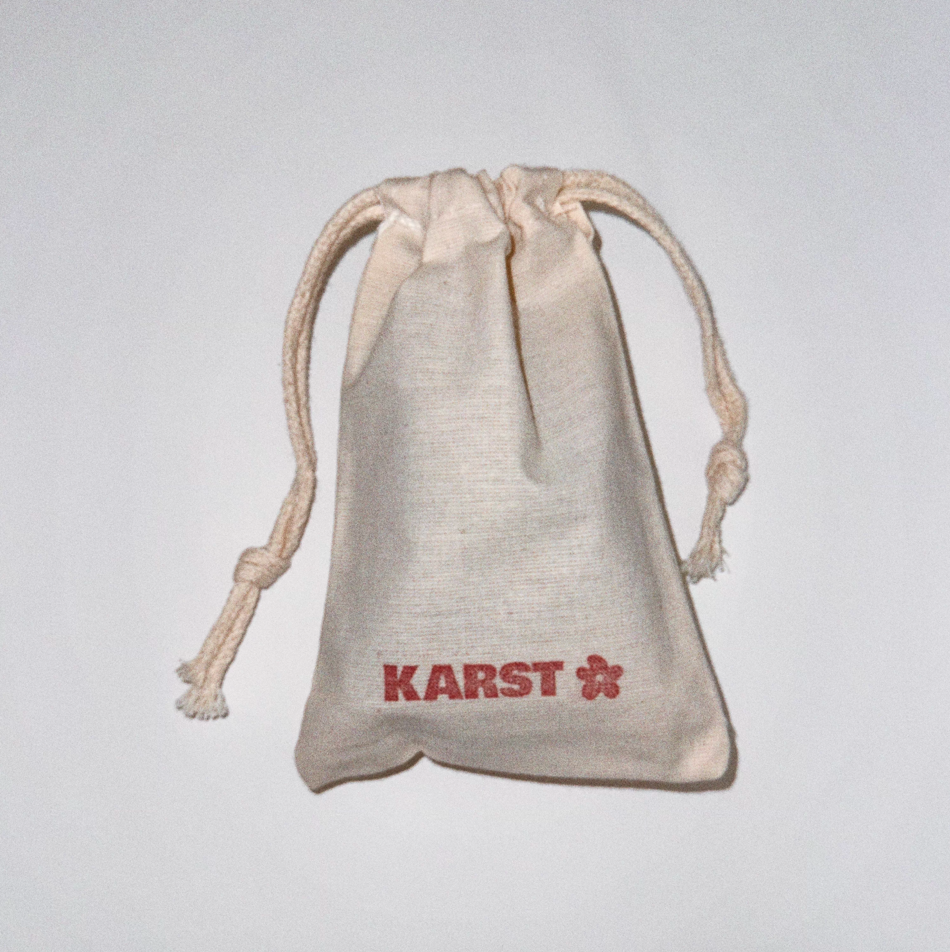 A small beige drawstring pouch with "Karst Essentials" and a red flower symbol printed on the front, perfect for storing your SuperClip Hair Claw — Buttermilk. Photographed against a plain white background.