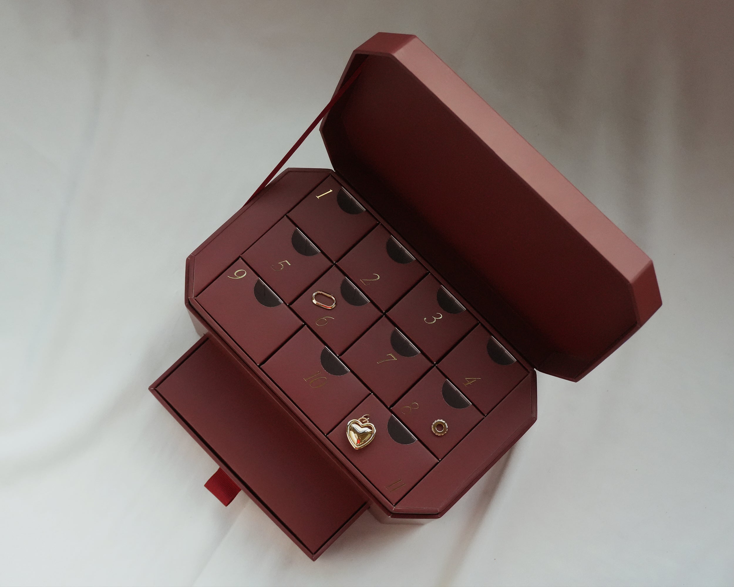 A maroon box with an open lid reveals twelve numbered drawers. Some drawers are partially open, displaying gold jewelry pieces, including a heart-shaped locket and a ring. The box sits on a white surface.
