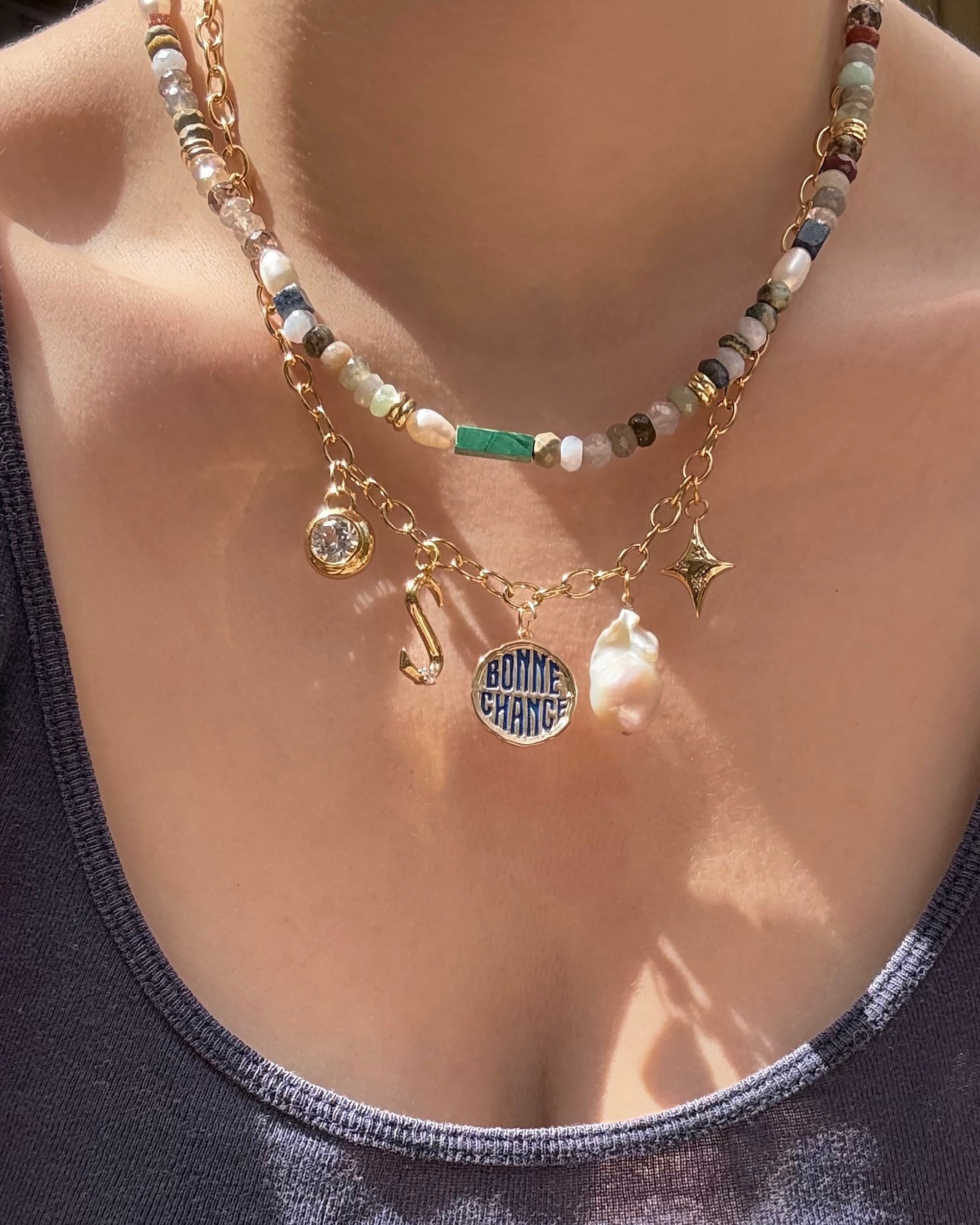 A close-up of a person wearing two layered Mod + Jo necklaces: one with colorful beads and another 14k gold chain featuring five charms—including a pearl, star, Tove Sapphire Bezel Charm, crystal, and round BONNE CHANCE pendant. Worn with gray tank top.