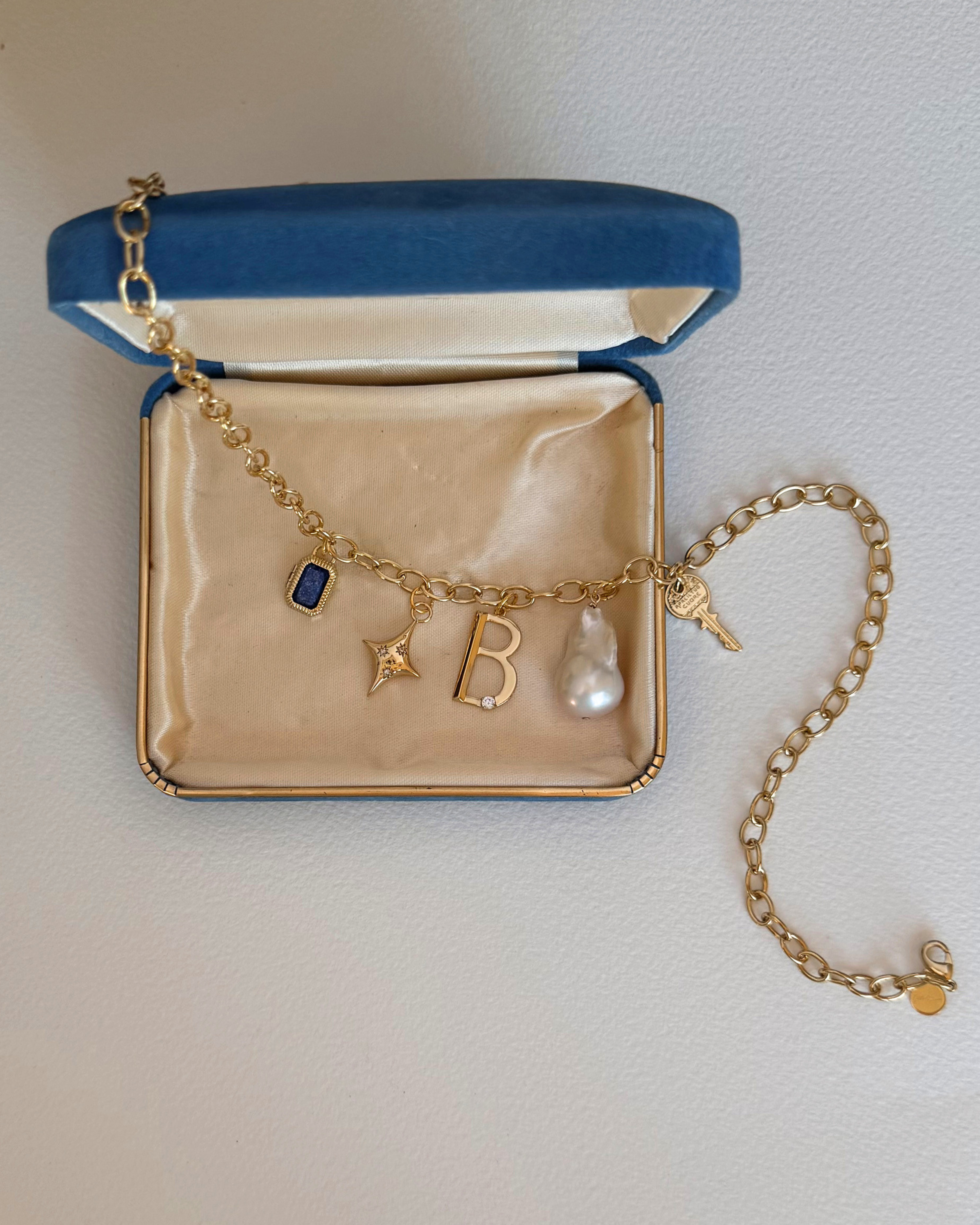 A Mod + Jo gold charm bracelet featuring the Celeste Puffy Star Charm, blue gem, letter "B," pearl, and key, displayed in an open blue velvet jewelry box with a cream-colored interior.