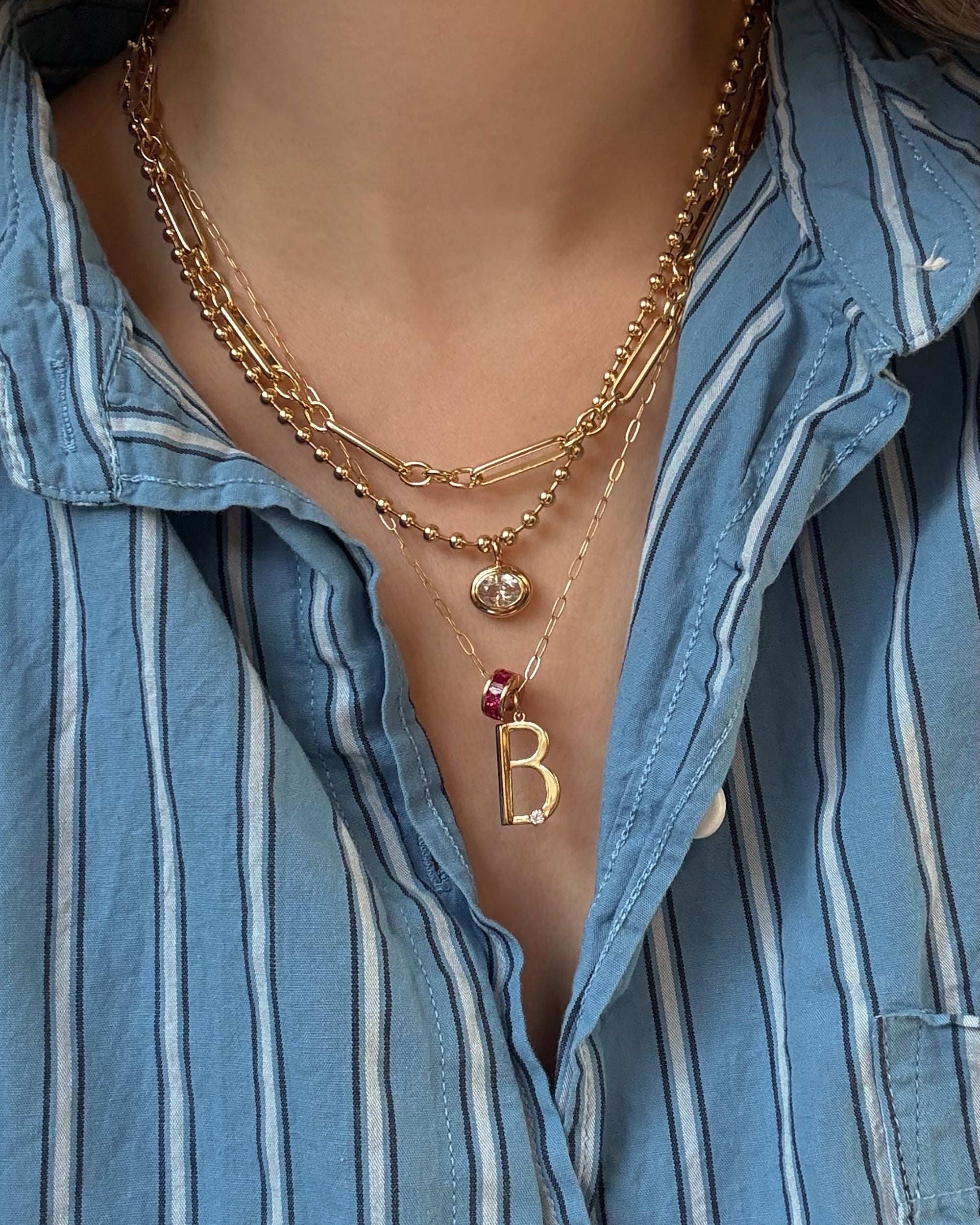 A person wearing a blue and white striped shirt and three layered gold necklaces, including Mod + Jo’s Eloise Letter Charm pendant with the letter B accented by small gems, and a gold sphere pendant.