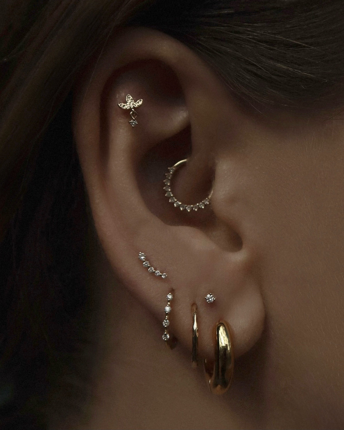 A close-up of an ear with multiple piercings, featuring a variety of gold and diamond earrings, including hoops, a curved bar, a butterfly stud, and a small diamond stud.
