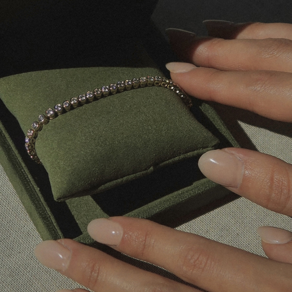 A person with manicured nails opens a green velvet jewelry box containing a delicate, gold and diamond tennis bracelet resting on a matching green cushion.