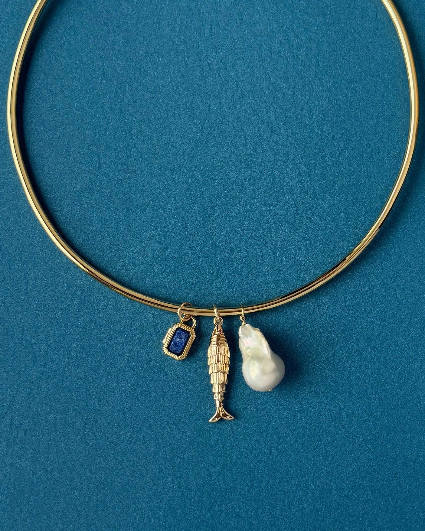 The Mod + Jo Jaci Baroque Pearl Charm is a 14k gold-plated bangle featuring three charms—a blue rectangular gem, a gold fish, and a white baroque pearl—displayed on a blue background.