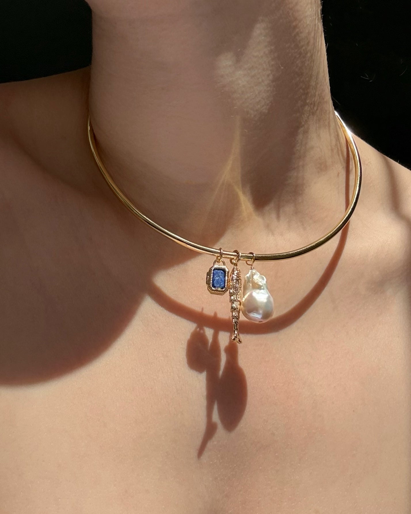 A close-up of the Mod + Jo Mabel Rectangle Birthstone Locket Charm, a gold choker necklace with interchangeable gemstone charms, worn on a neck as sunlight casts shadows on the skin.