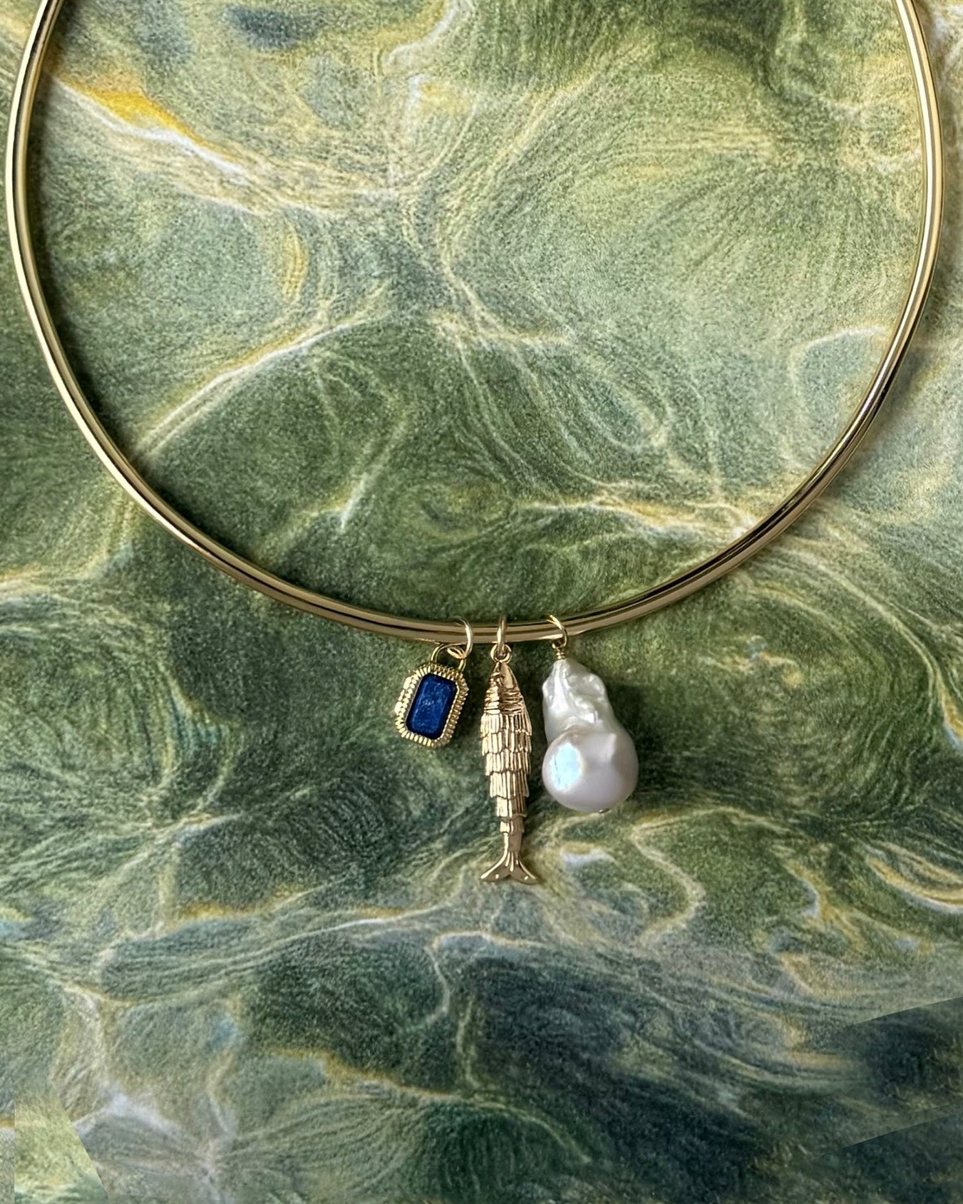 The Mod + Jo Mabel Rectangle Birthstone Locket Charm, a small blue gem, features on a gold bangle with a fish and large white baroque pearl, all displayed against a green marble-like background.