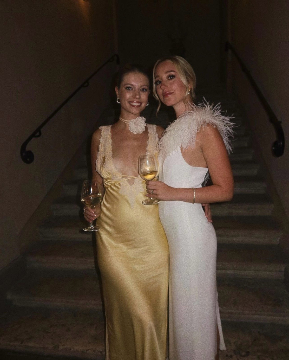 Two women in elegant evening gowns stand side by side on a staircase, each holding a glass of white wine and smiling at the camera. One wears a yellow dress, and the other wears a white dress with feather details.