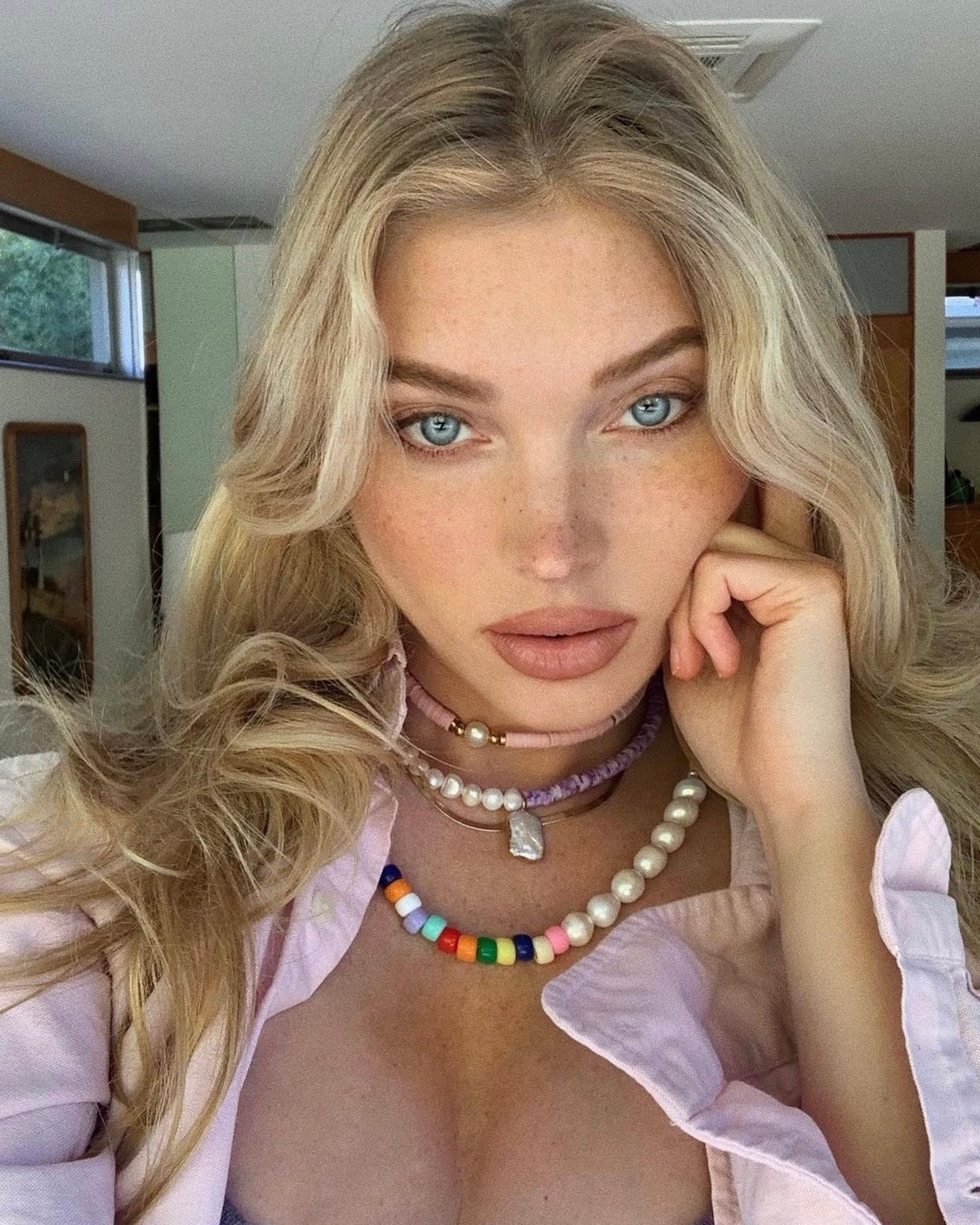 A woman with long blonde hair and blue eyes poses indoors, resting her head on her hand. She wears layered colorful bead and pearl necklaces, a light purple top, and a light pink shirt, with natural light illuminating her face.