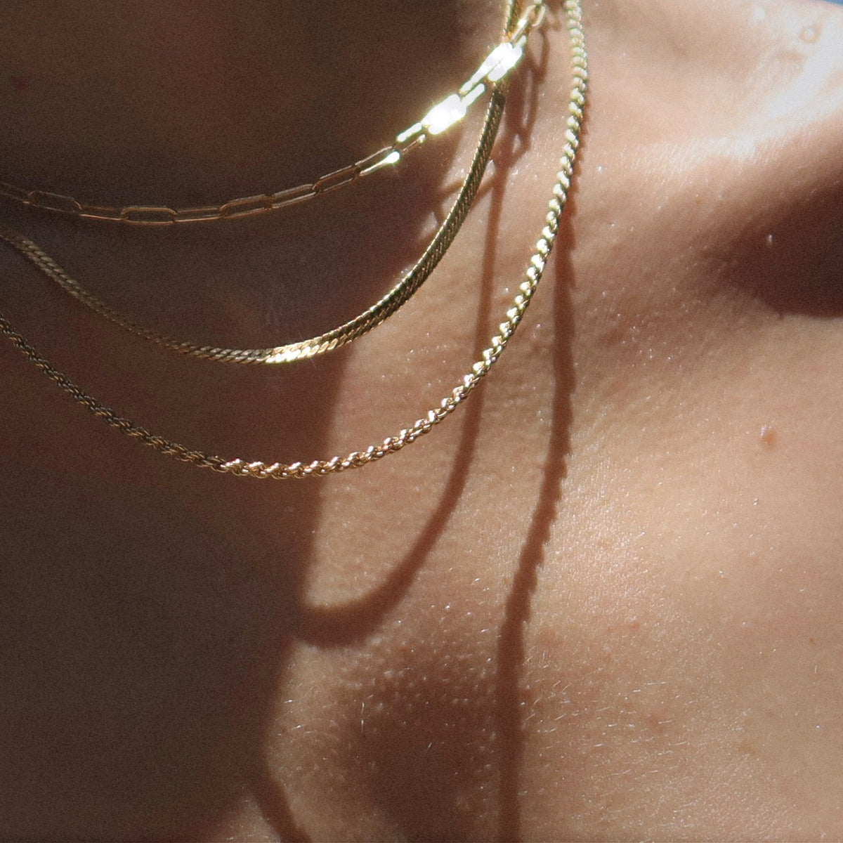 Close-up of a persons neck and collarbone in soft light, wearing three delicate gold necklaces with different chain styles. Shadows from the chains fall on the skin.