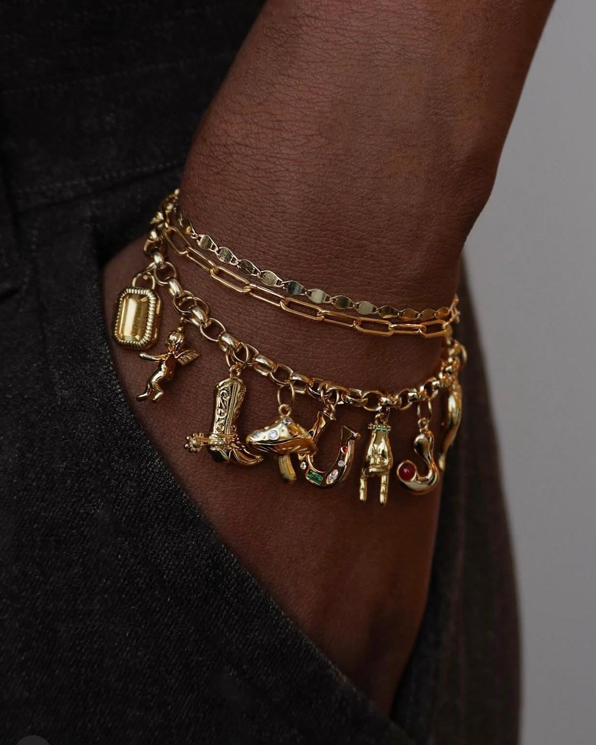 A close-up of a person’s wrist wearing layered gold chain bracelets with various quirky gold charms, including a boot, a shoe, a hand, a cow, and other playful shapes. The hand is in a dark pocket.