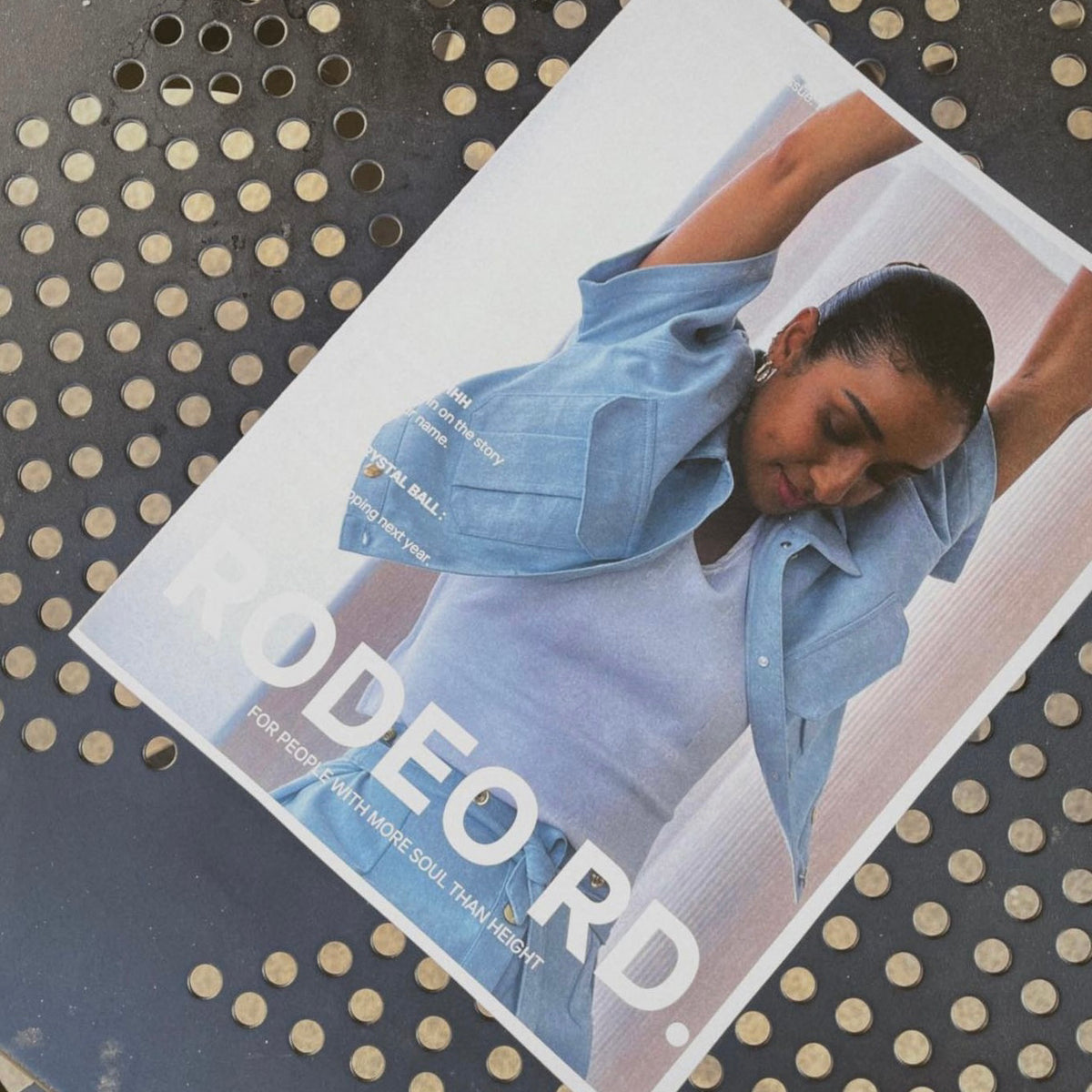 A magazine with a photo of a woman stretching her arms upward in a light blue shirt and white top lies on a perforated metal surface. The magazine cover reads RODEORD. in large white letters.