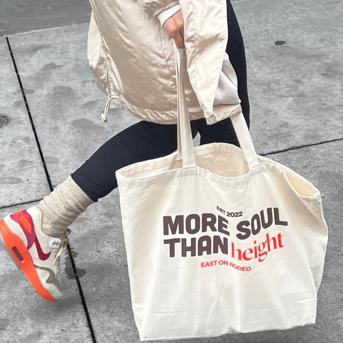 A person in a light coat and colorful sneakers walks on a sidewalk, carrying a beige tote bag that reads More Soul Than Height with height in red and EST 2022, East on Rodeo beneath.
