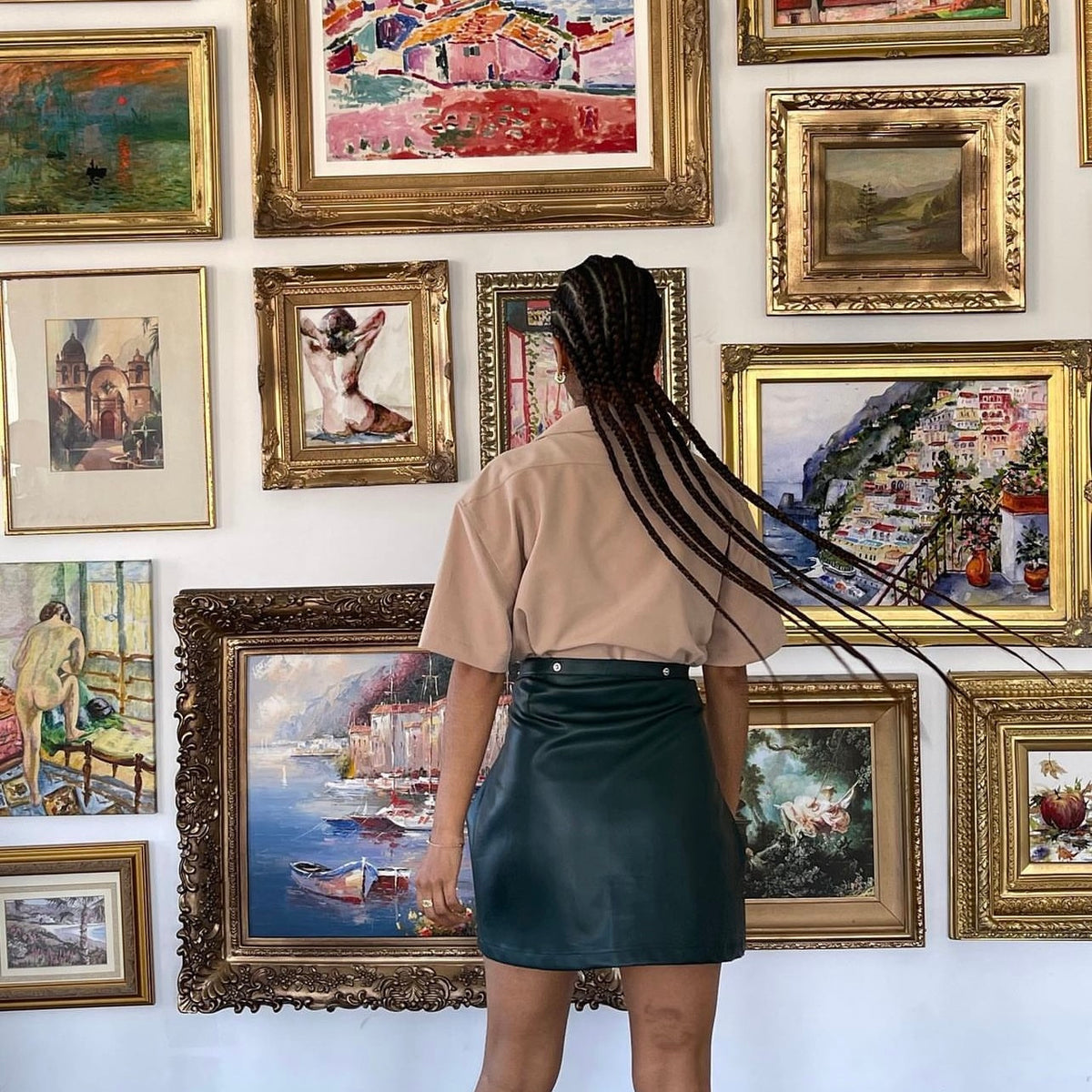 A woman with long braids, wearing a beige shirt and a dark green skirt, stands facing a wall filled with colorful framed paintings of various scenes and styles.