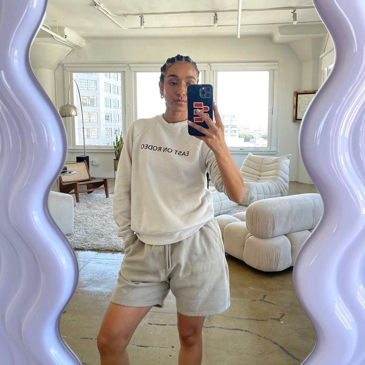 A woman stands in a modern, bright living room, taking a mirror selfie. She wears a white sweatshirt with EAST ON ARCO text and light gray shorts. The wavy-edged mirror frames her, and stylish furniture is visible behind her.