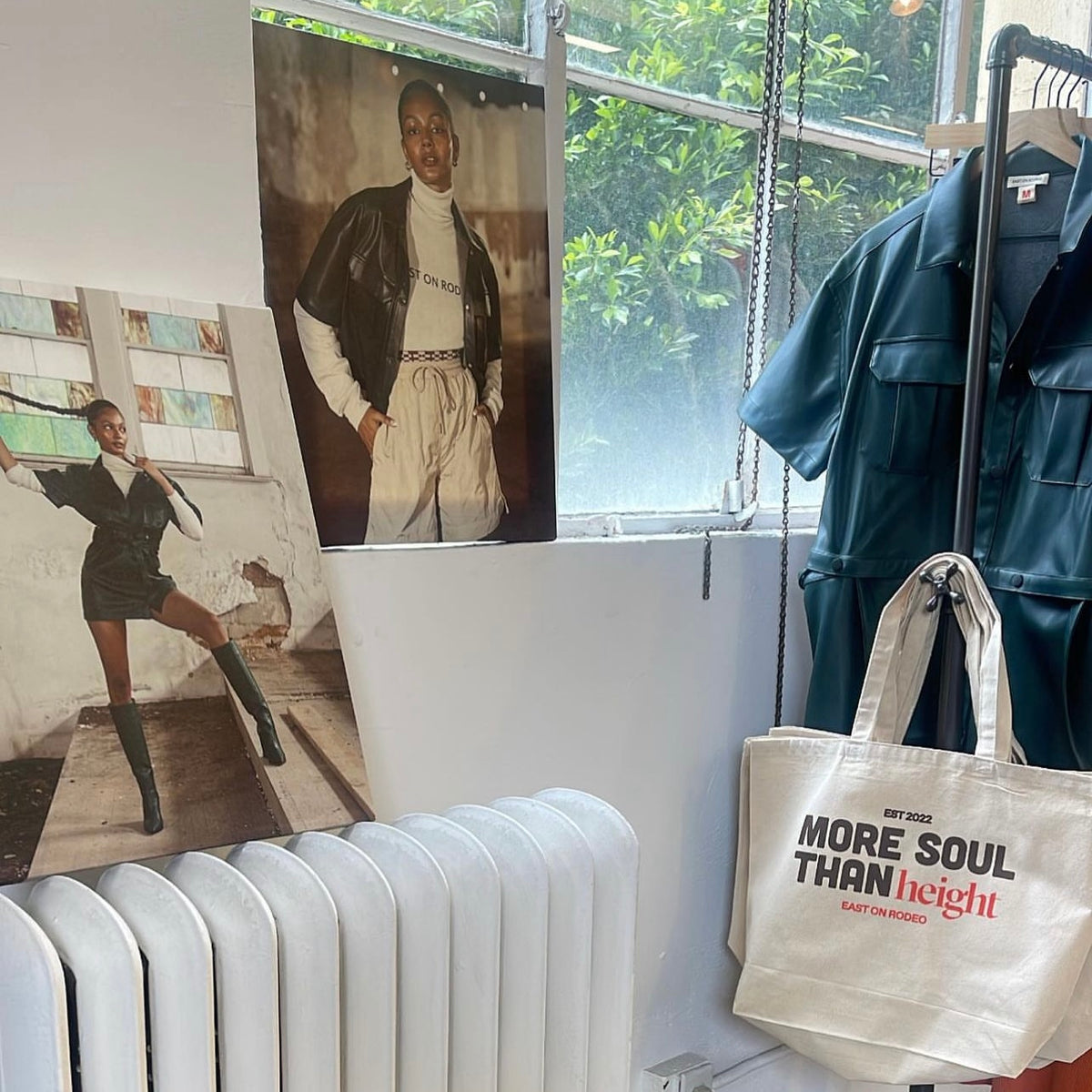 Two fashion posters of a model, a blue faux-leather outfit on a hanger, and a white tote bag reading MORE SOUL THAN height are displayed in a sunlit room with a radiator and window.