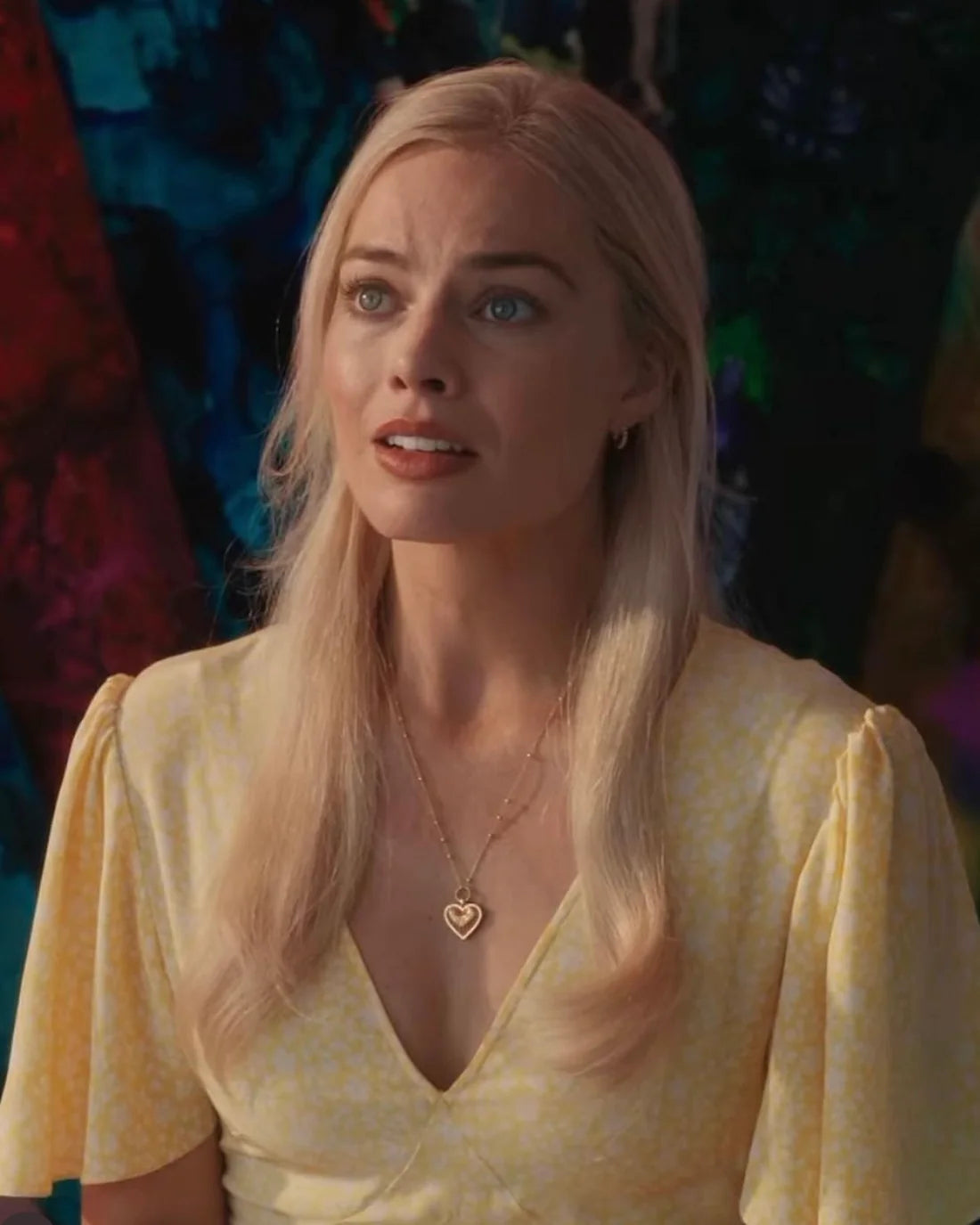 A woman with long blonde hair wearing a light yellow dress and a heart-shaped necklace looks slightly upward with a concerned expression. The background is colorful and abstract.