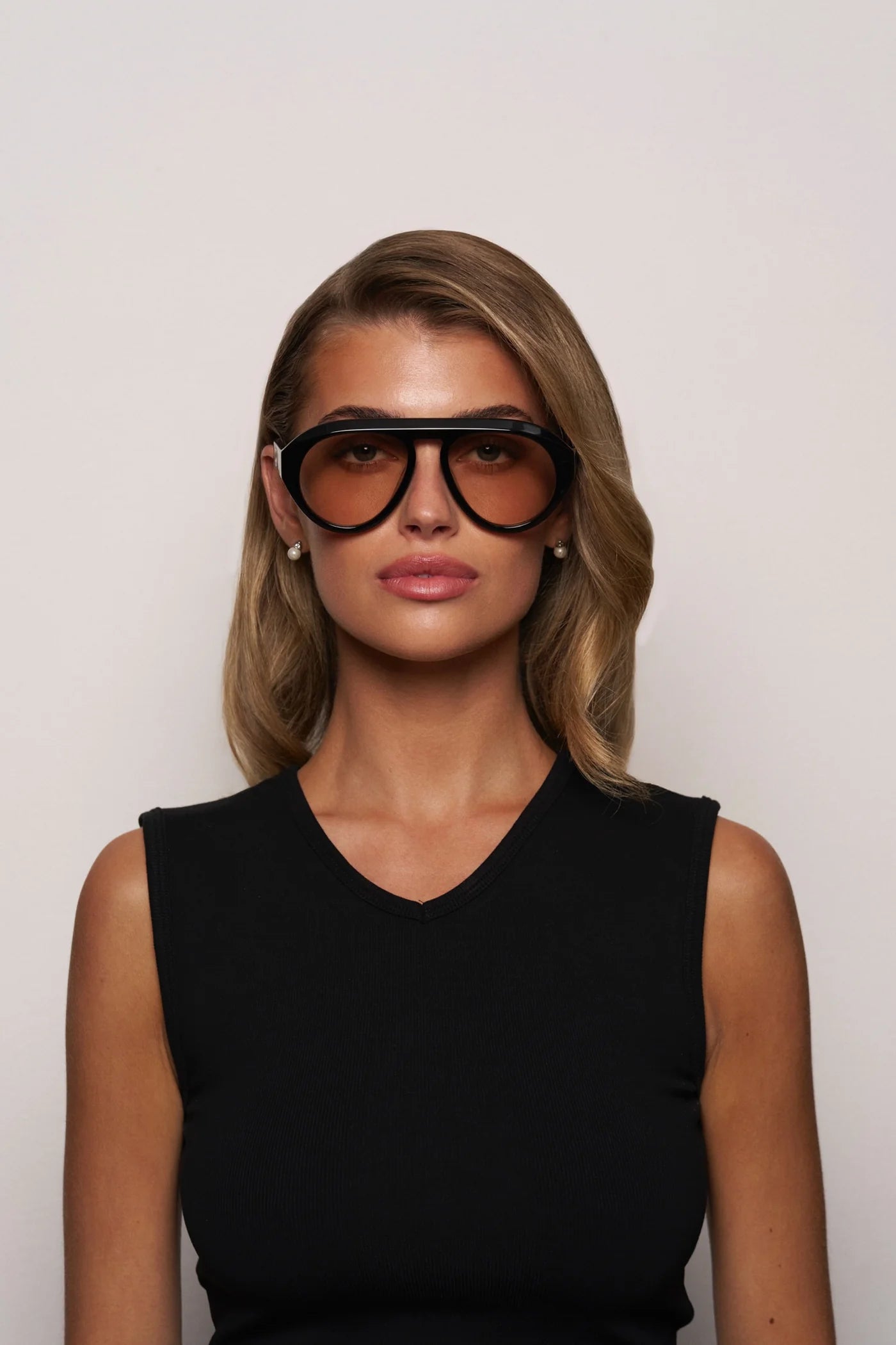 A woman with wavy blonde hair wears Elisa Johnson's Val Sunglasses, large acetate aviators with UV400 protection, and a sleeveless black top, posing against a plain light background.