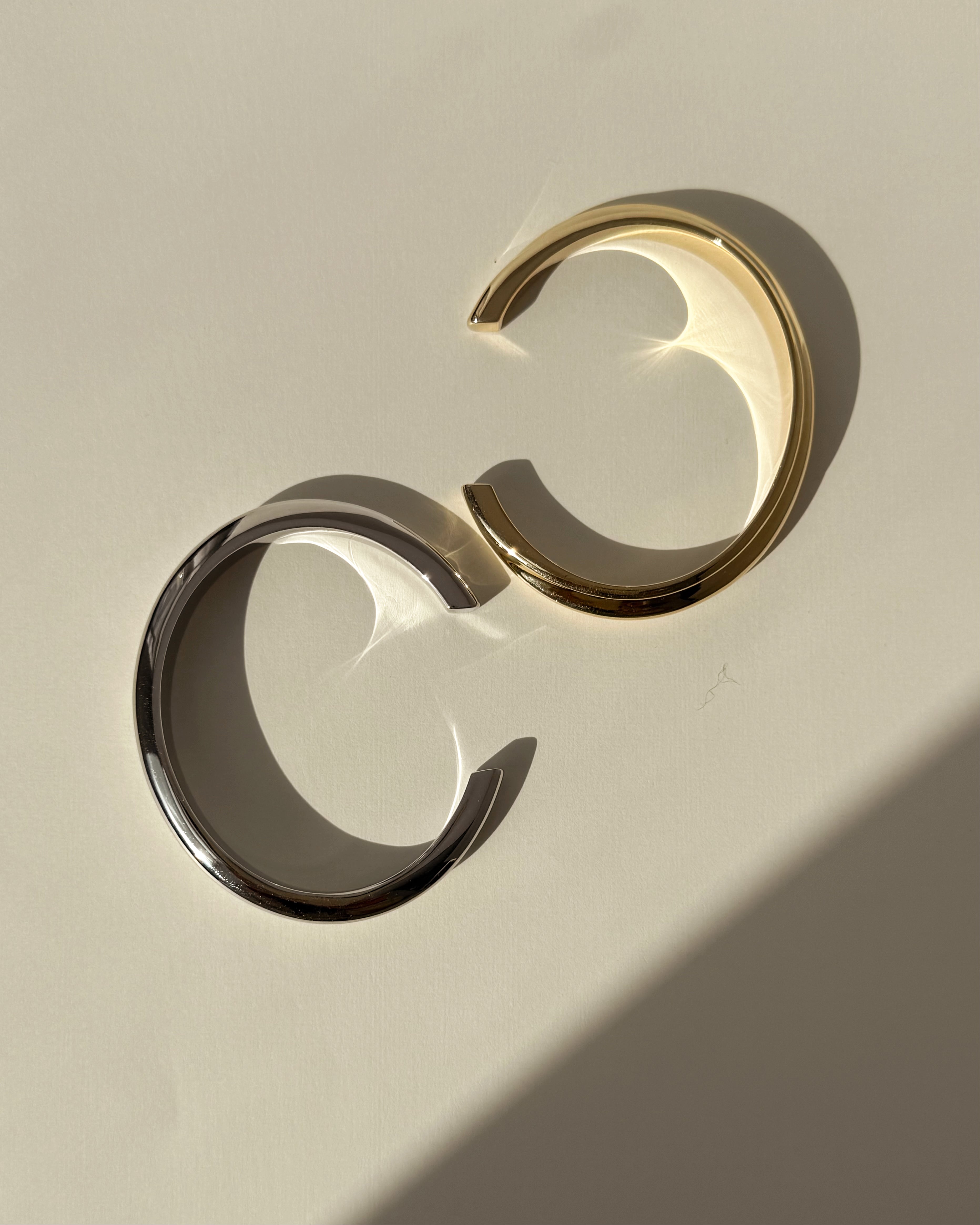 Two Leila Dome Cuff Bracelets by Mod + Jo—one gold plated, one silver—rest on a light surface. Gentle shadows and reflections highlight their elegant design, ideal for any 14k gold jewelry collection.