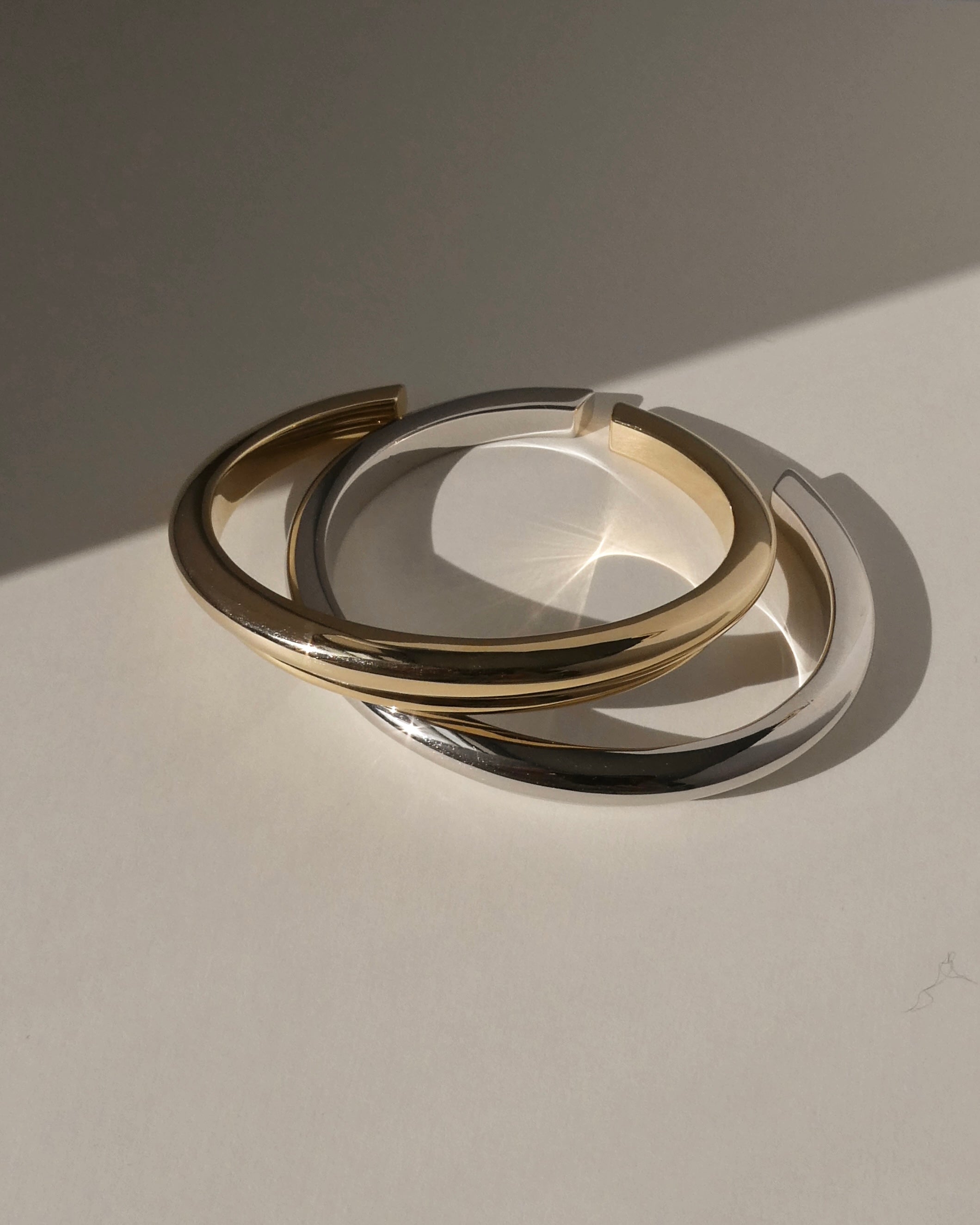 Two minimalist Leila Dome Cuff Bracelets by Mod + Jo—one in polished gold and one in silver—overlap on a smooth, light surface, casting soft shadows and reflections in natural sunlight.