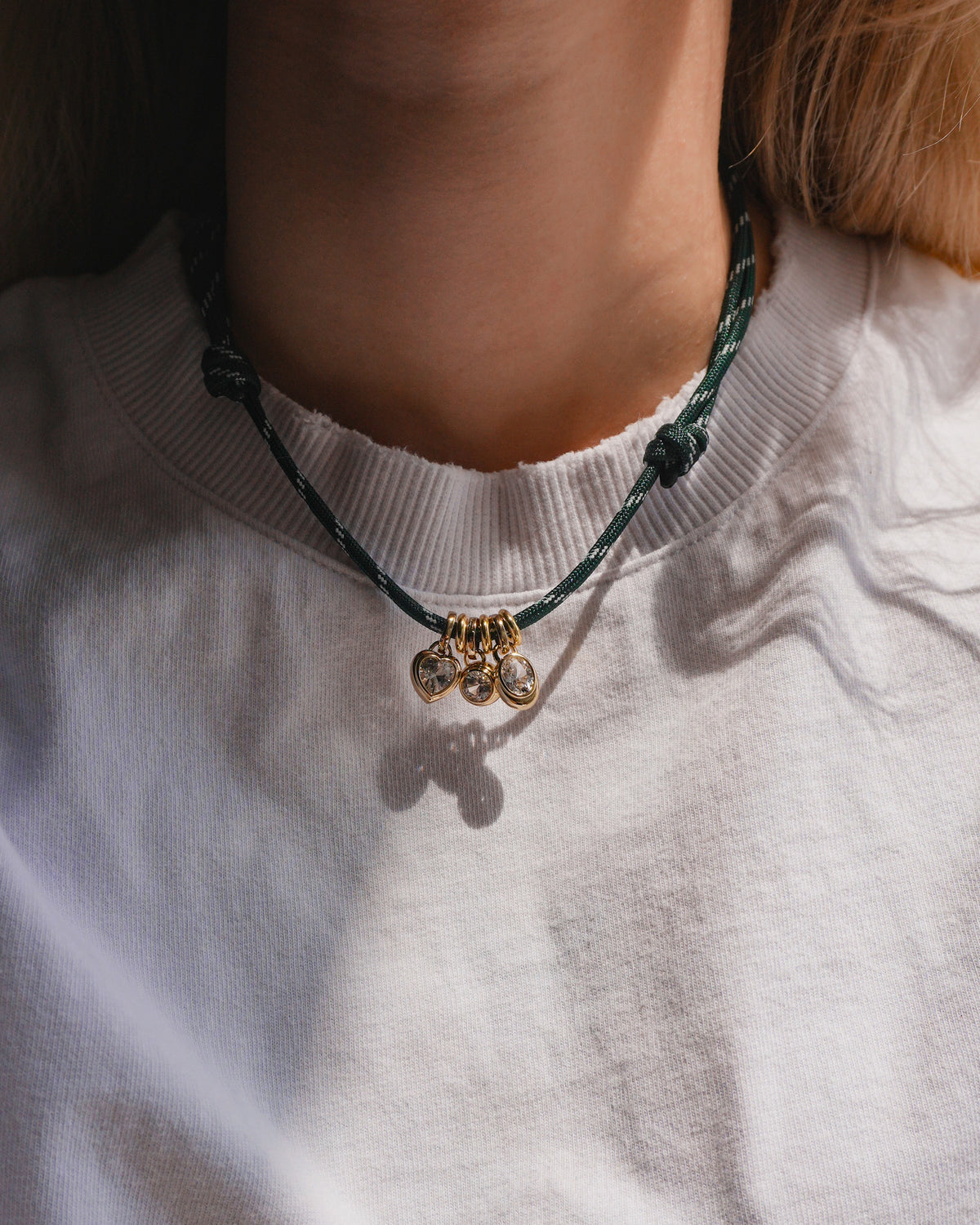 A person wearing a white crewneck shirt and a Mod + Jo Tommy Cord Necklace—featuring a green paracord, three small shiny gold pendant charms, and gold-plated spacers—is photographed at chest level.