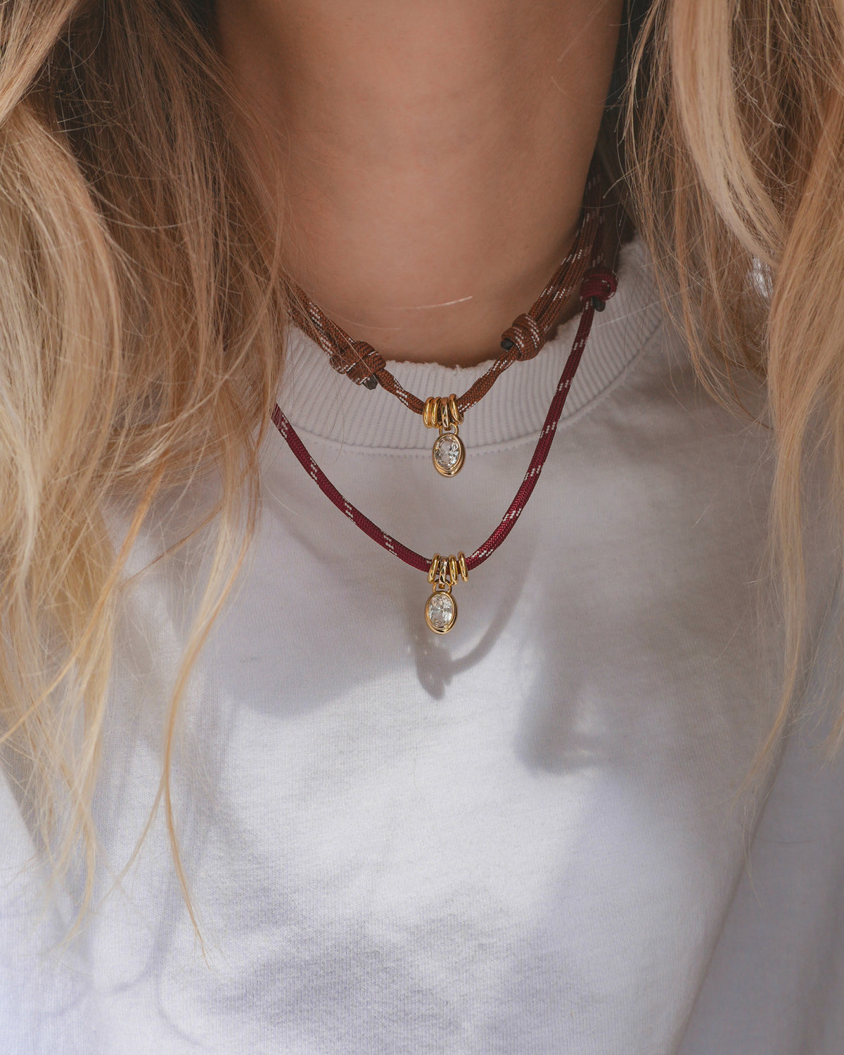 A person wearing a white top and two necklaces—one is the Mod + Jo Tommy Cord Necklace, featuring gold and diamond pendants with gold-plated spacers on adjustable red and brown cords. Their light brown hair partially frames the image.