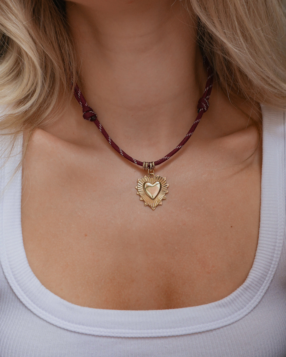 A woman wearing a white ribbed tank top and the Mod + Jo Tommy Cord Necklace—a maroon paracord necklace featuring gold plate accents and a gold heart-shaped pendant; only her neck, collarbone, and part of her hair are visible.