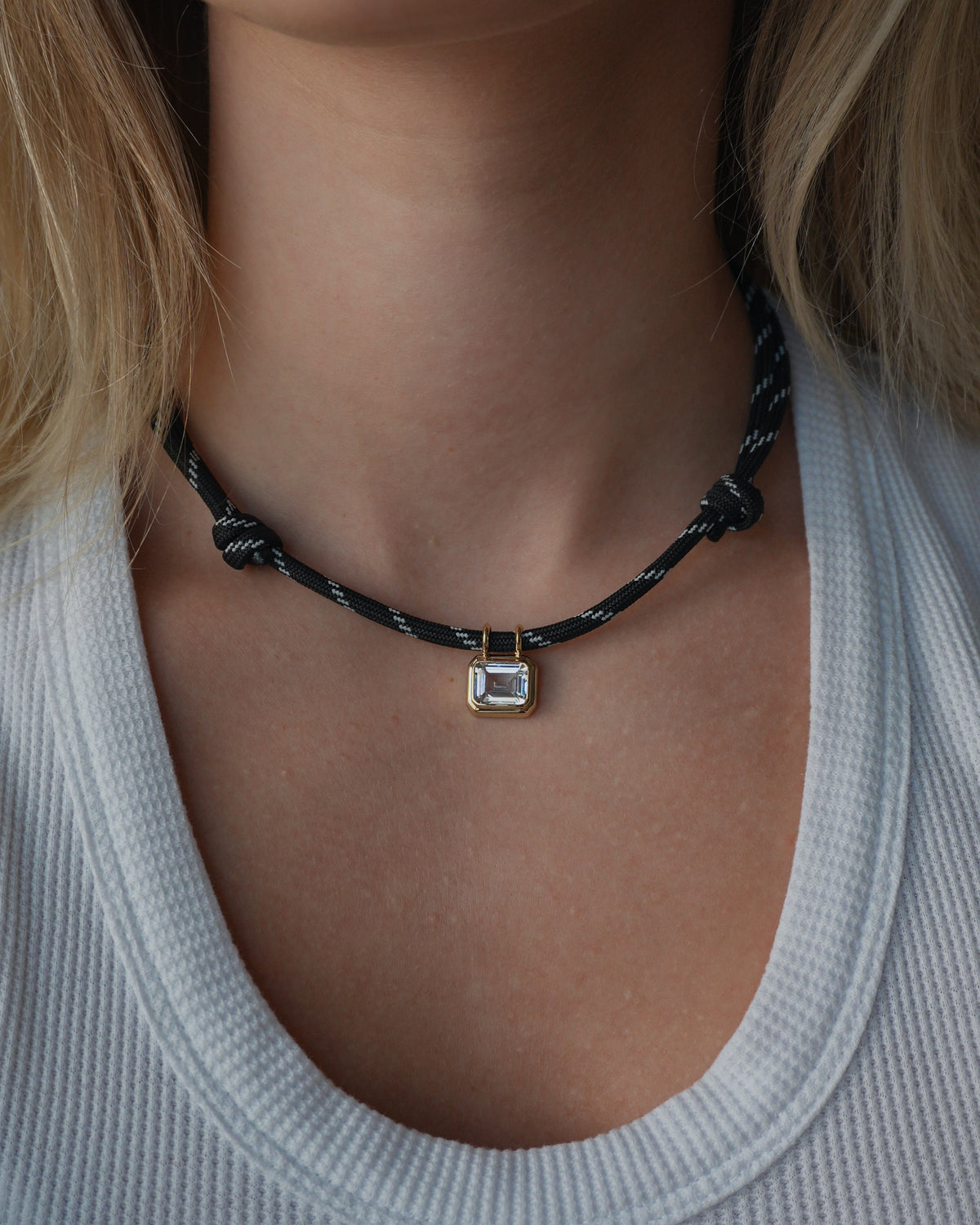 A close-up of a person wearing a white textured shirt and the Mod + Jo Tommy Sapphire Pendant Necklace, featuring a black adjustable paracord and a rectangular pendant set with a lab-grown white sapphire in gold.