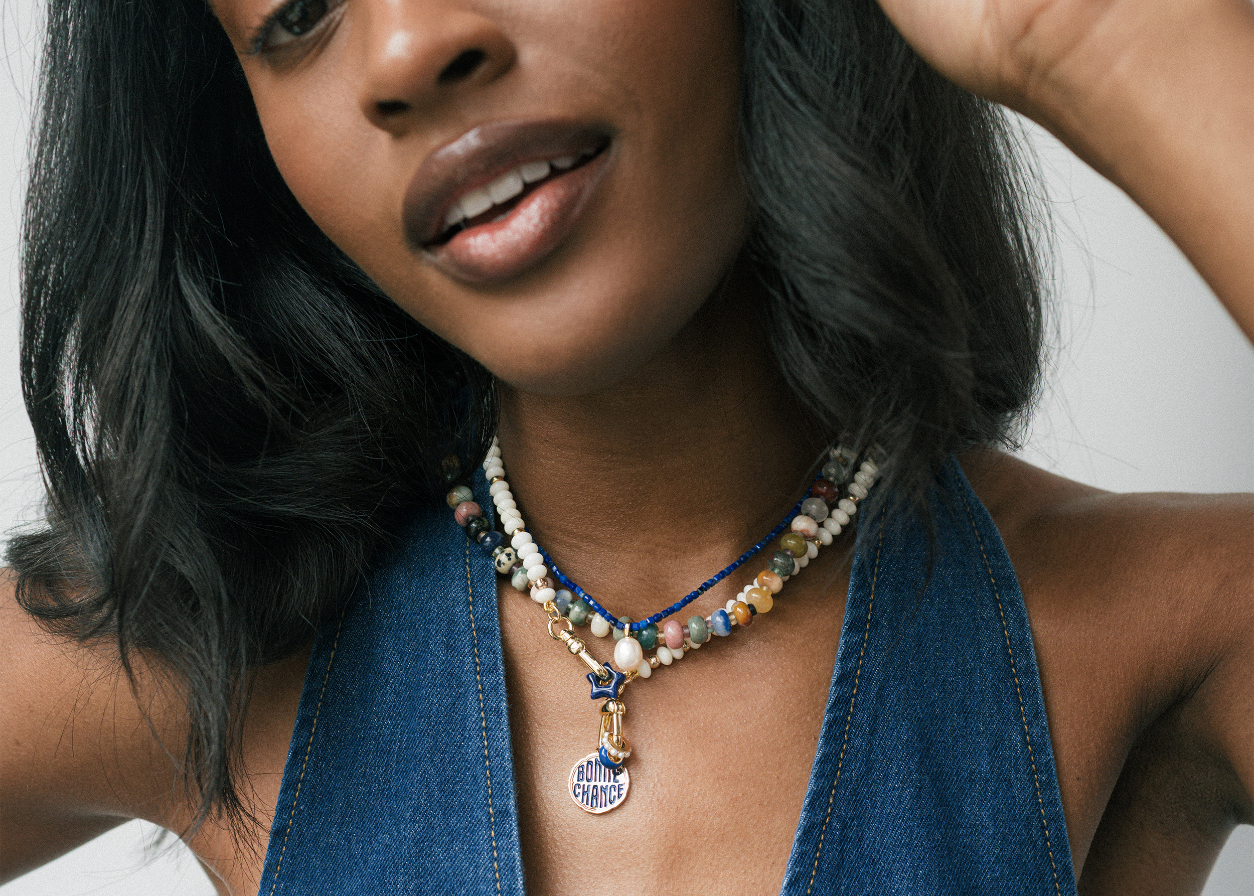A woman wearing a sleeveless denim top and layered beaded necklaces, including one with a round FOR CHANGE pendant, smiles softly with her head slightly tilted.