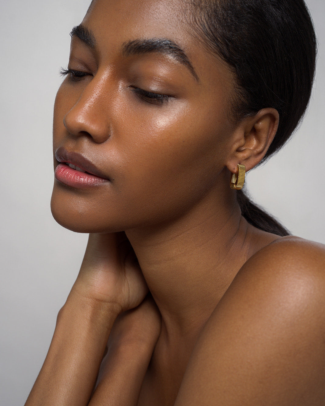 A woman with smooth, glowing skin poses with her eyes closed, hand gently touching her neck. She wears the Elly Fluted Hoops by Mod + Jo—lightweight gold earrings that complement her pulled-back dark hair. The background is plain and light-colored.