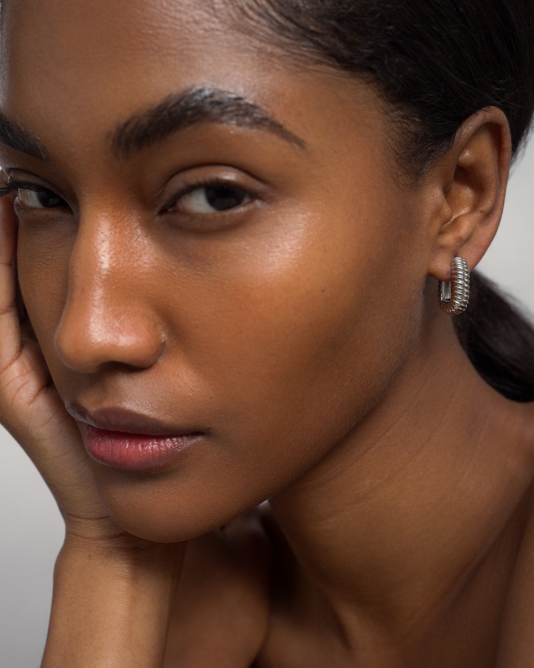 A close-up of a woman with smooth brown skin, dark hair pulled back, and arched eyebrows, elegantly wearing the Mod + Jo Elly French Rope Hoops and gently resting her hand on her face.