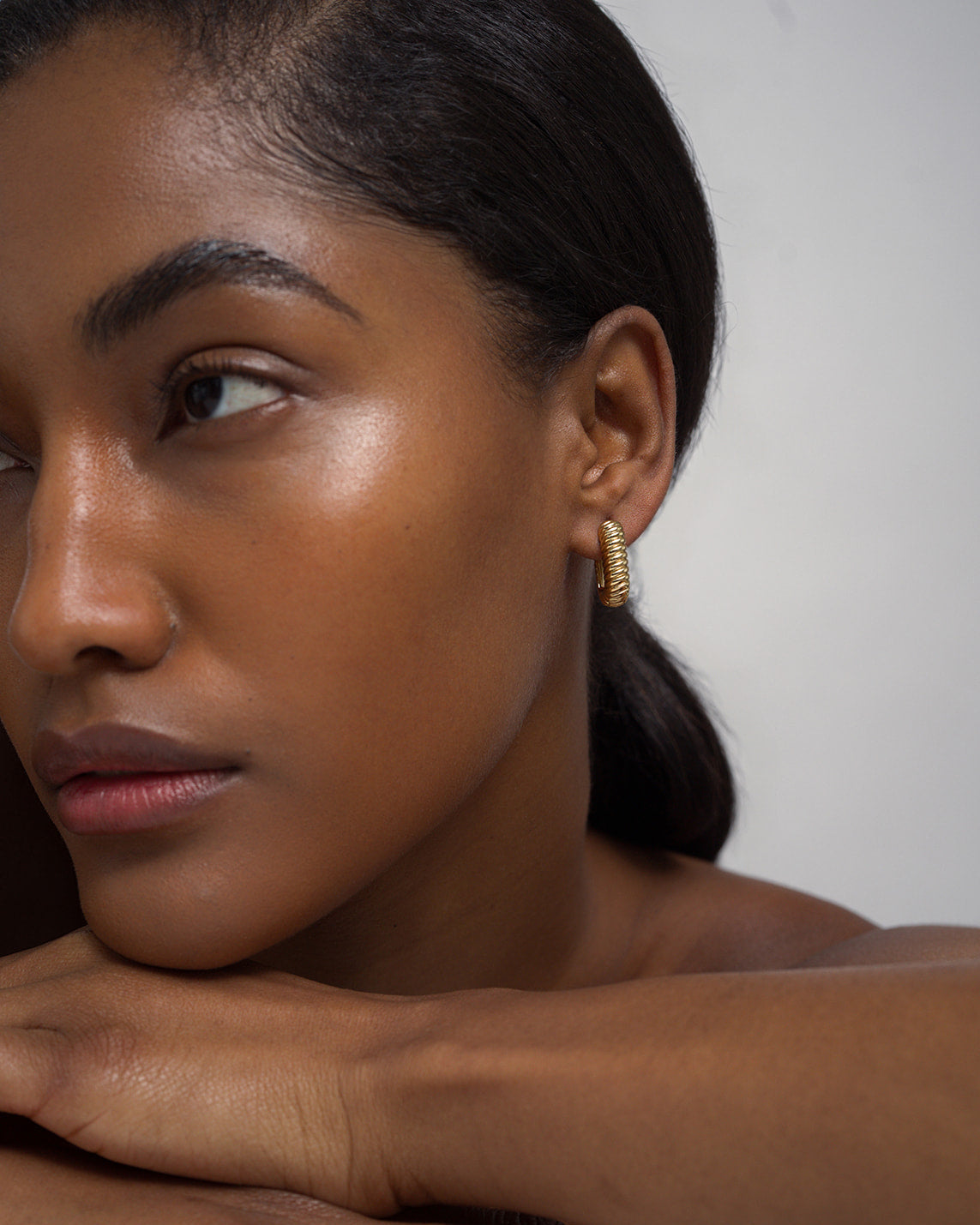 A woman with smooth, glowing skin rests her chin on her arms, gazing to the left. Her dark hair is pulled back, and she wears the Mod + Jo Elly French Rope Hoops—their textured gold design elegantly catching the light. The background is plain and light.