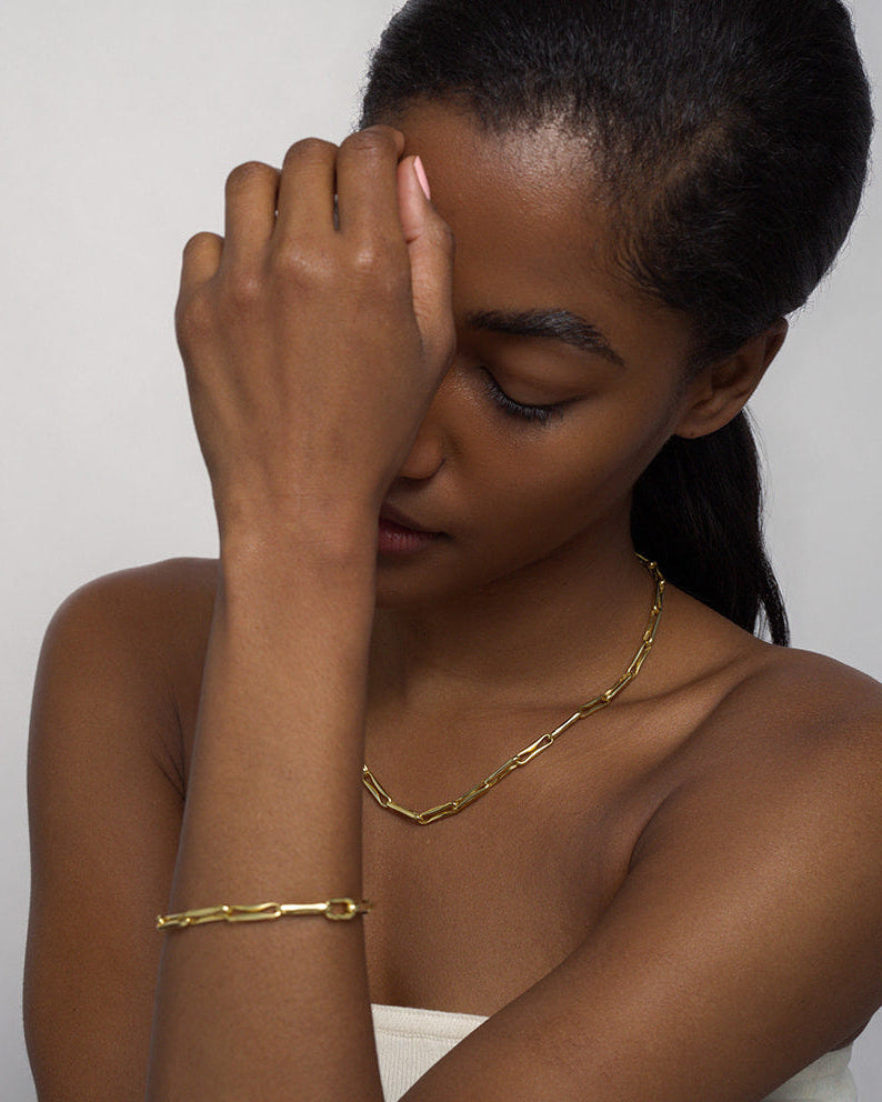 A woman with dark hair and smooth skin poses with her arm partially covering her face, wearing a gold chain necklace and the Mod + Jo Molly Elongated Link Chain Bracelet against a neutral background.