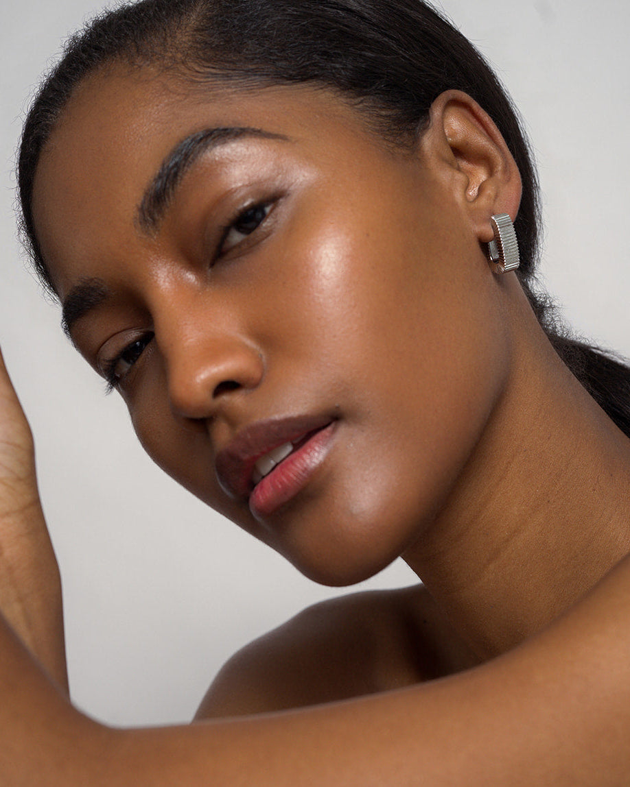A woman with smooth, glowing skin poses against a light background, wearing the Elly Fluted Hoops by Mod + Jo. Her dark hair is pulled back as she gazes softly at the camera with a relaxed expression.