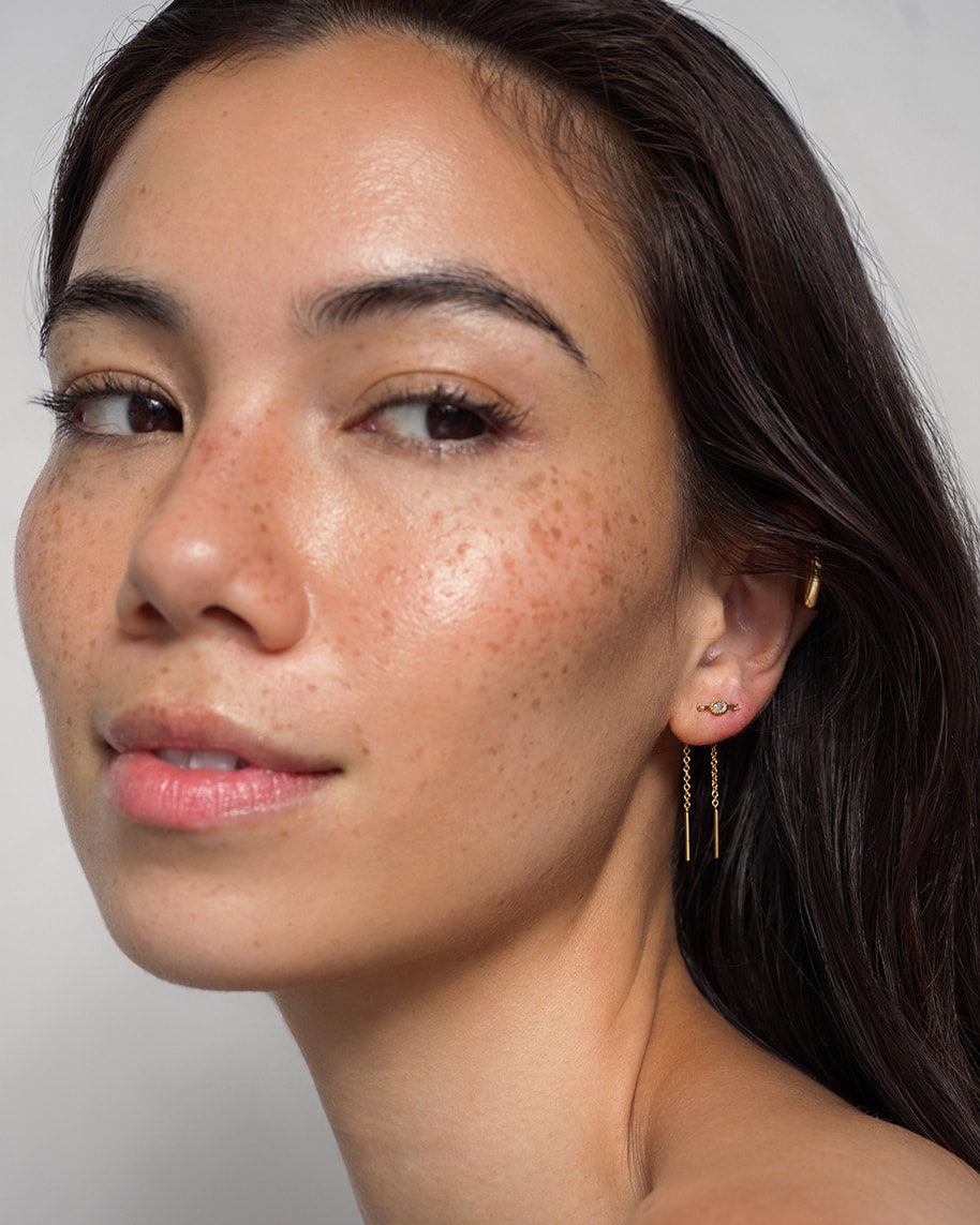 A woman with long dark hair and freckles gazes at the camera, wearing minimal makeup and the Mod + Jo Nicole Oval Threader Earrings, elegantly plated in gold. The background is light and neutral.
