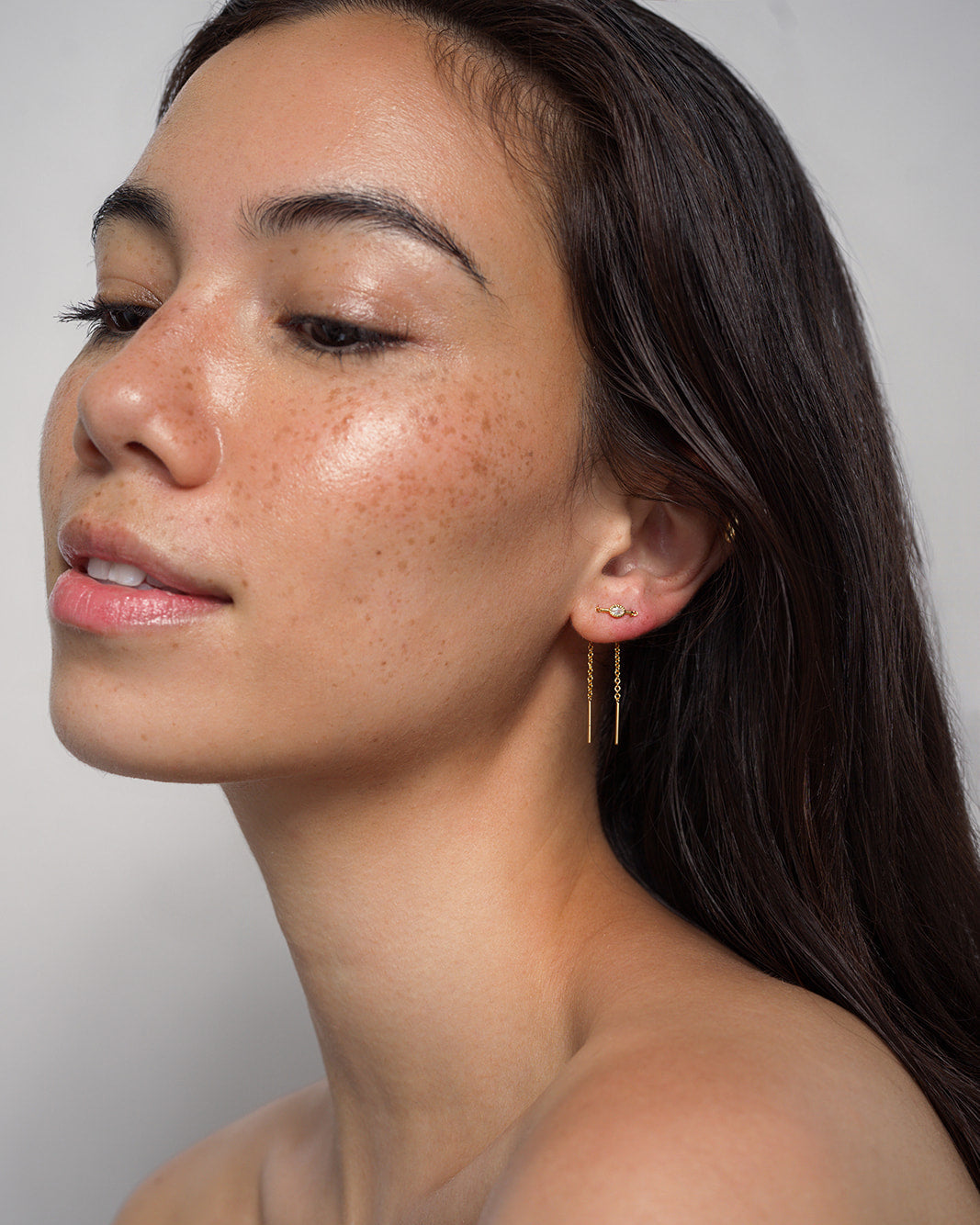A woman with long dark hair and freckled skin looks to the side, wearing the Mod + Jo Nicole Oval Threader Earrings. Her complexion is dewy and she wears minimal makeup against a neutral background.