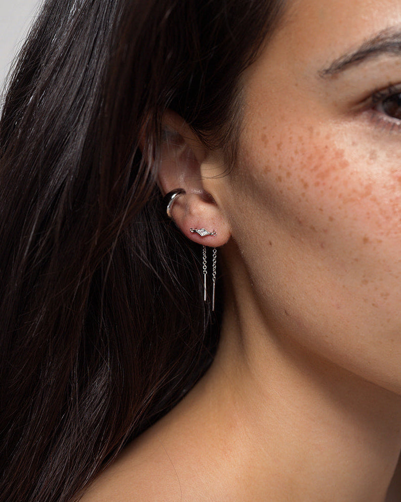 Close-up of a woman’s ear adorned with three earrings: a small hoop, a cubic zirconia stud with a curved design, and the Mod + Jo Nicole Kite Threader Earring. She has long dark hair, freckles, and light brown skin. The image focuses on her rhodium-plated jewelry.