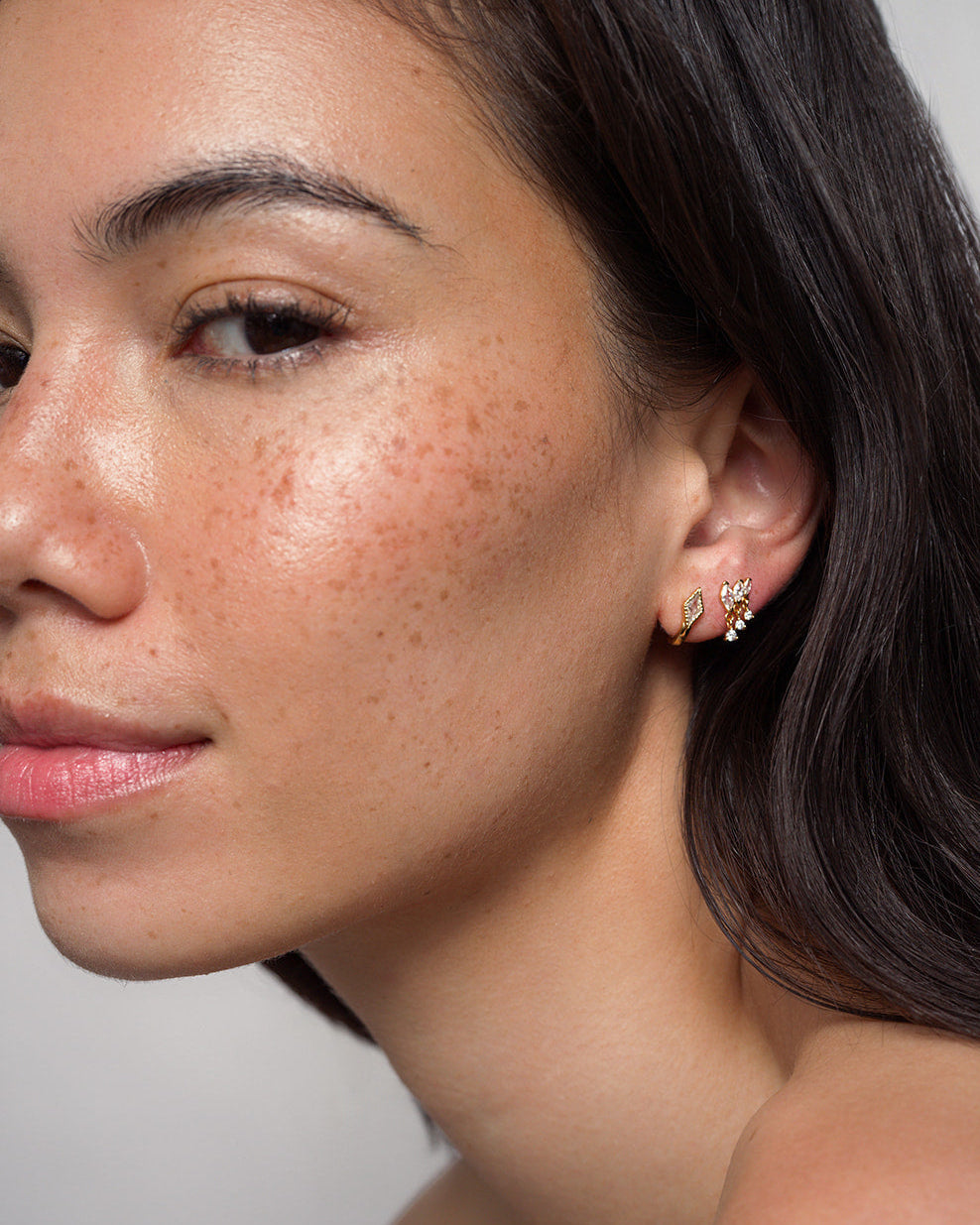 A woman with smooth, freckled skin and long dark hair wears the Gabby Marquise Dangle Studs by Mod + Jo—two small, gold-plated earrings delicately shaped like leaves. She gazes softly at the camera against a plain background.