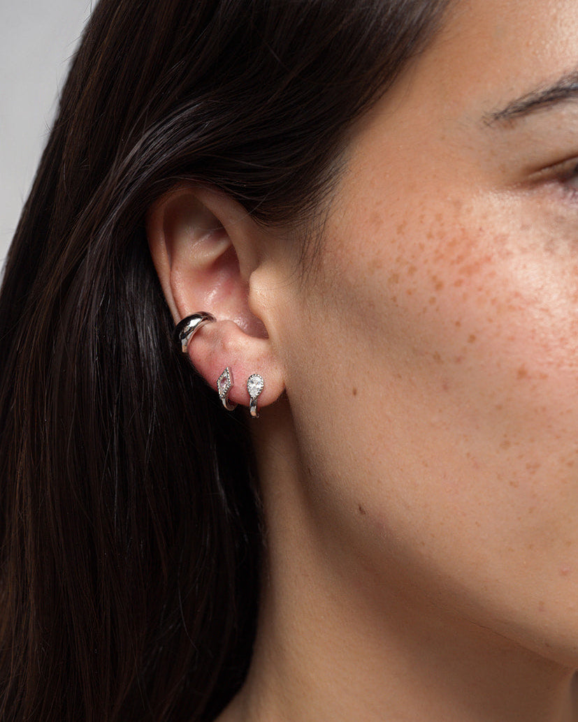 A close-up shows a woman with dark hair and light, freckled skin wearing four silver earrings from Mod + Jo. At the top of her ear, she wears a rhodium-plated hoop. On her lower ear are three Leighton Teardrop Huggies—small, leaf-shaped studs adorned with clear cubic zirconia gemstones.